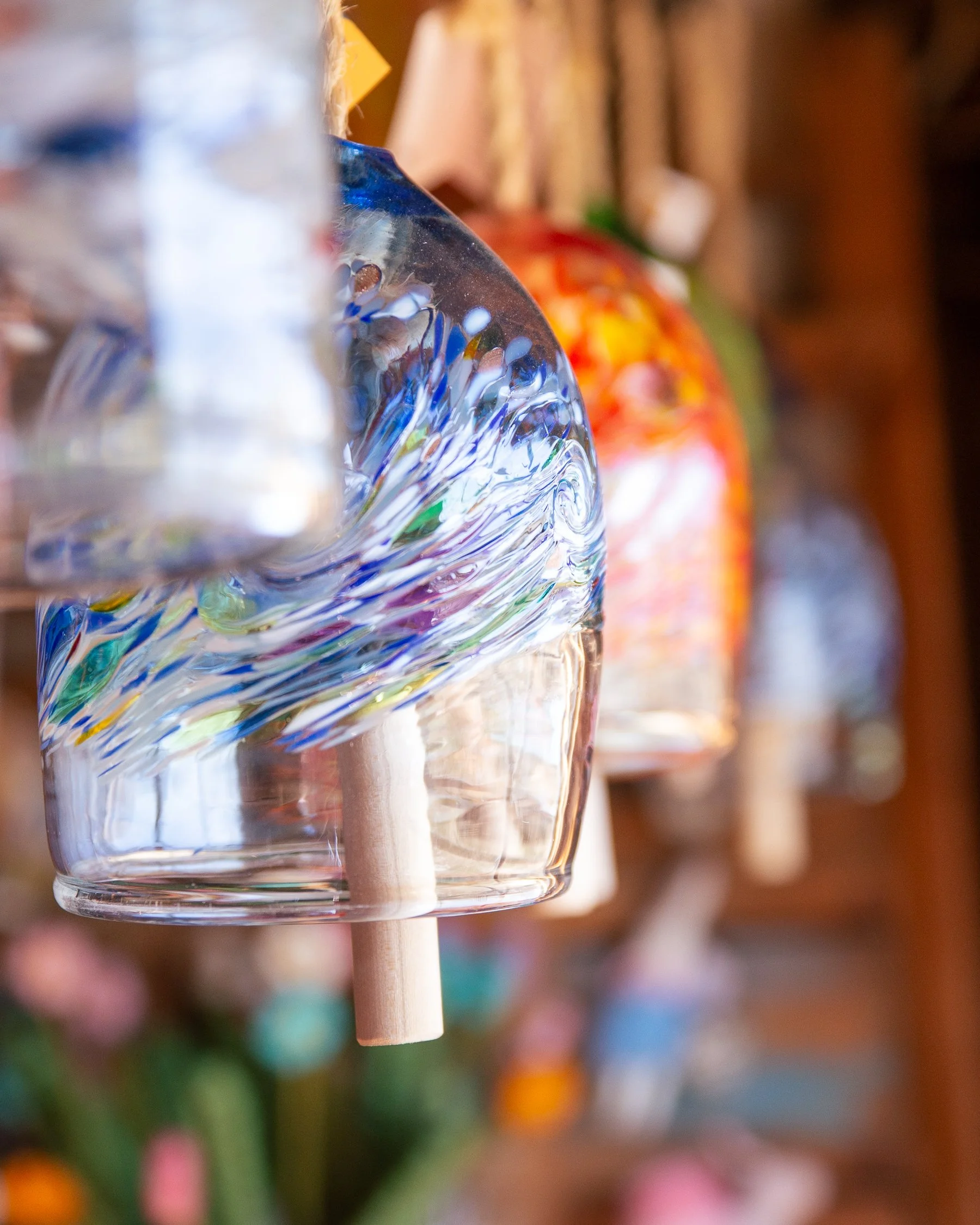 Close-up of a colorful glass wind chime bell with swirls of blue, purple, green, and yellow, with wooden clapper, with blurred multicolored wind chimes or ornaments in the background.