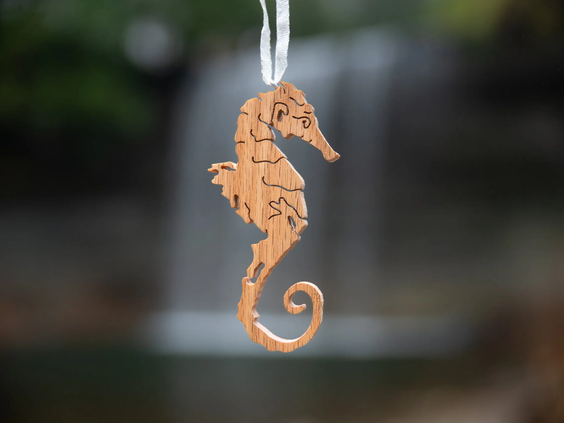 Wooden seahorse ornament that includes outlines of Manitoulin Island hanging by a white ribbon, with a blurred natural waterfall background.