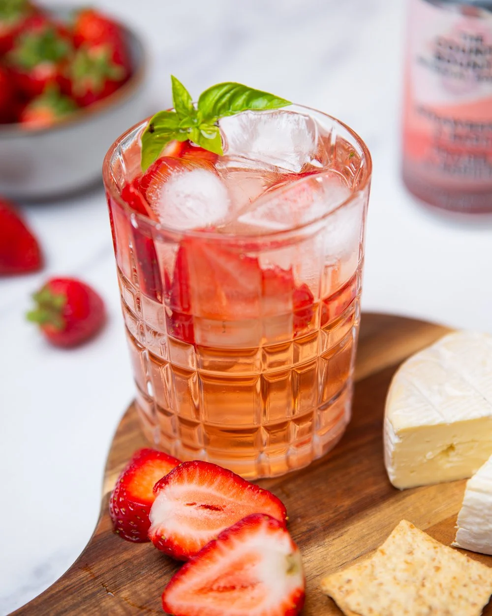 A glass of strawberry cocktail with ice and fresh basil garnish on a wooden serving board, surrounded by strawberries, cheese, and crackers, with a bowl of strawberries and can of The County Bounty strawberry basil artisanal soda in the background