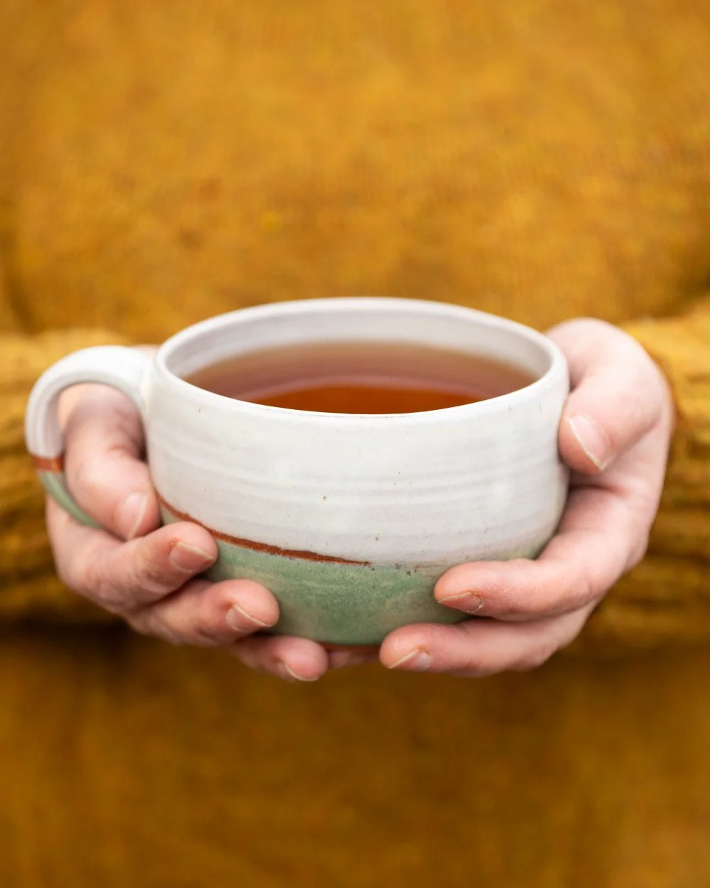 Person holding a white and teal dual-toned ceramic cup filled with tea, wearing a yellow knitted sweater.