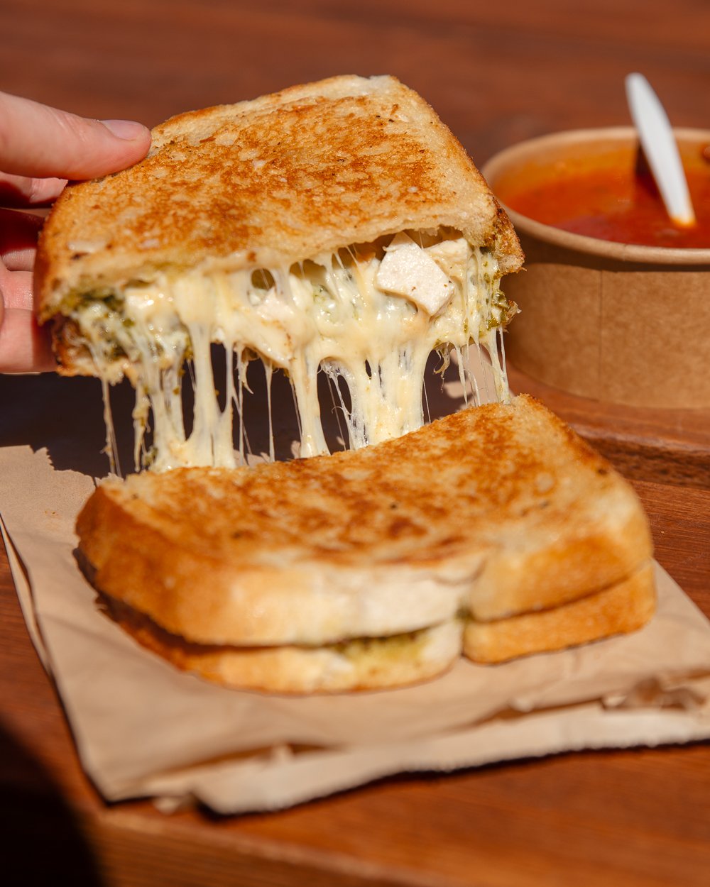 Close-up of a grilled cheese sandwich with gooey, melted cheese, being lifted to show the melty interior, containing what looks to be pesto and chicken, with a bowl of tomato soup in the background.