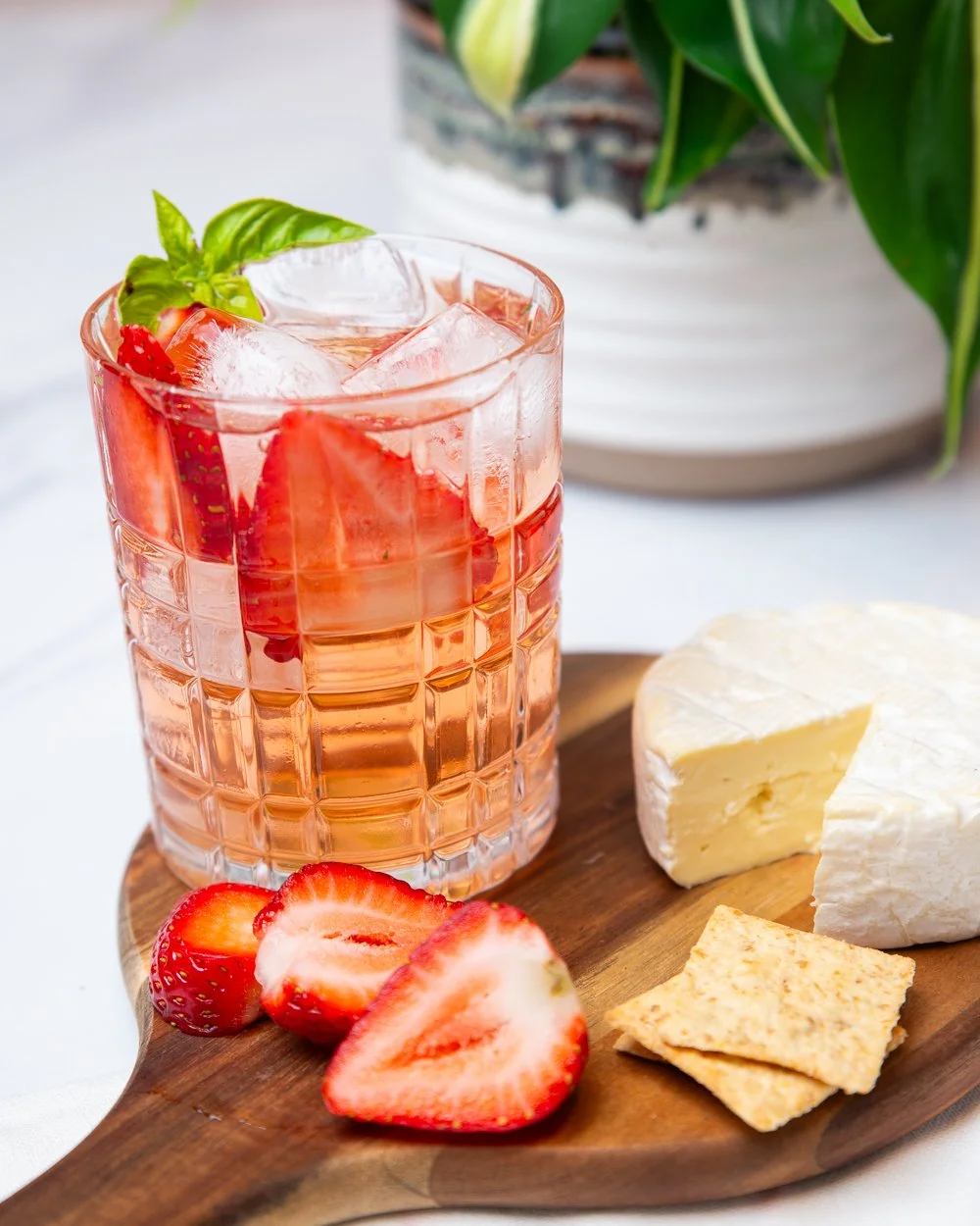 A glass of pink strawberry lemonade with ice, garnished with fresh strawberries and basil, placed on a wooden serving board alongside a wedge of brie cheese, crackers, and sliced strawberries, with a potted green plant in the background.