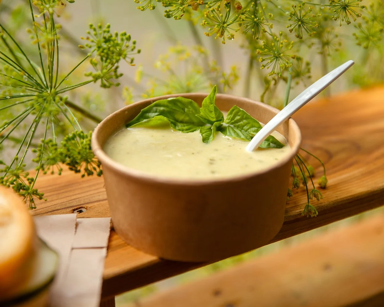Bowl of creamy green vegetable soup garnished with basil leaves and a white spoon, placed on a wooden surface with green dill plants in the background.