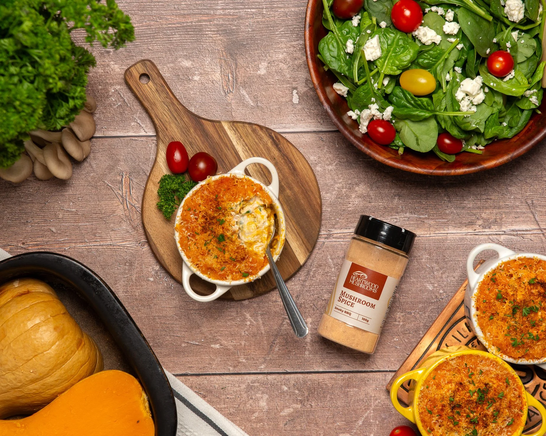 A wooden table with a large salad bowl filled with leafy greens, cherry tomatoes, and crumbled feta cheese. There are small baking dishes with cheesy baked dishes, and a jar of mushroom spice. A part of a squash and a soup spoon are also visible.