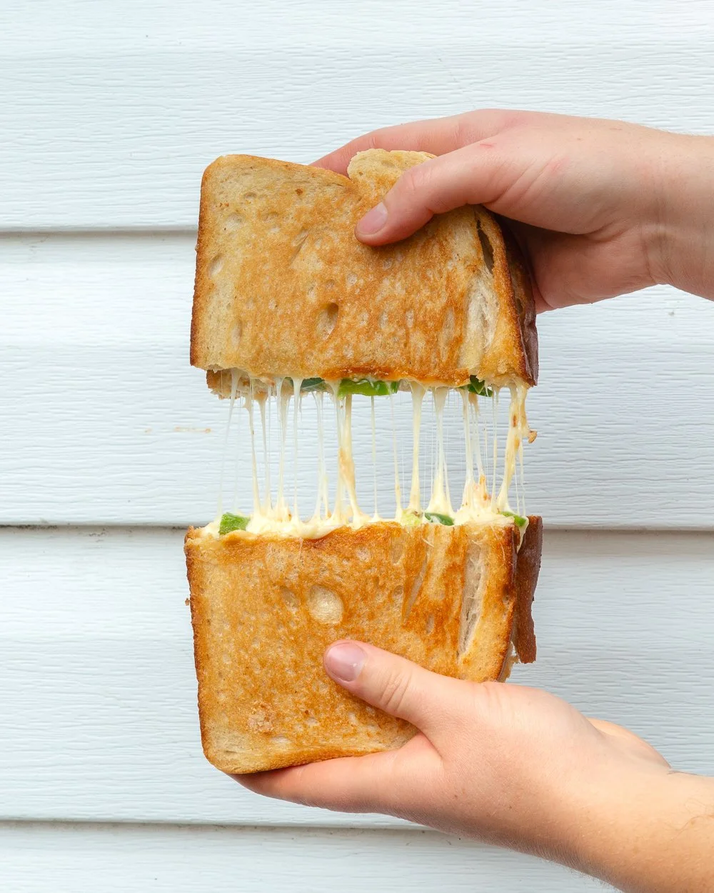 Person holding a grilled cheese sandwich with melted cheese stretching between the bread slices, against a white wooden background.