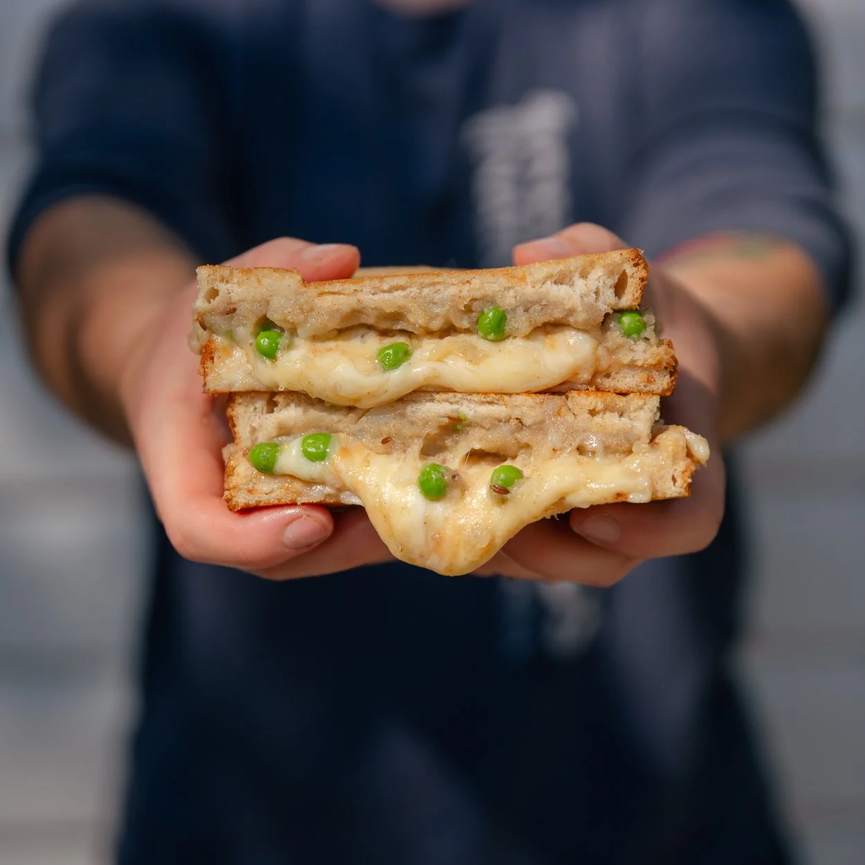 Person holding a sandwich with melted cheese, green peas, and toasted bread slices.