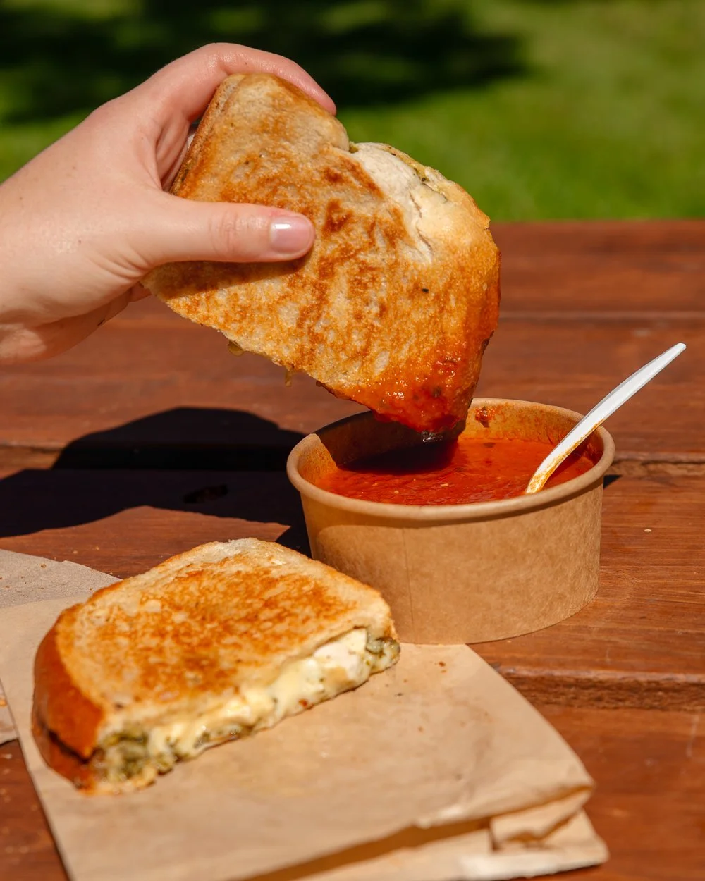 Hand dipping a slice of grilled cheese sandwich into a bowl of tomato soup on a wooden table outdoors.