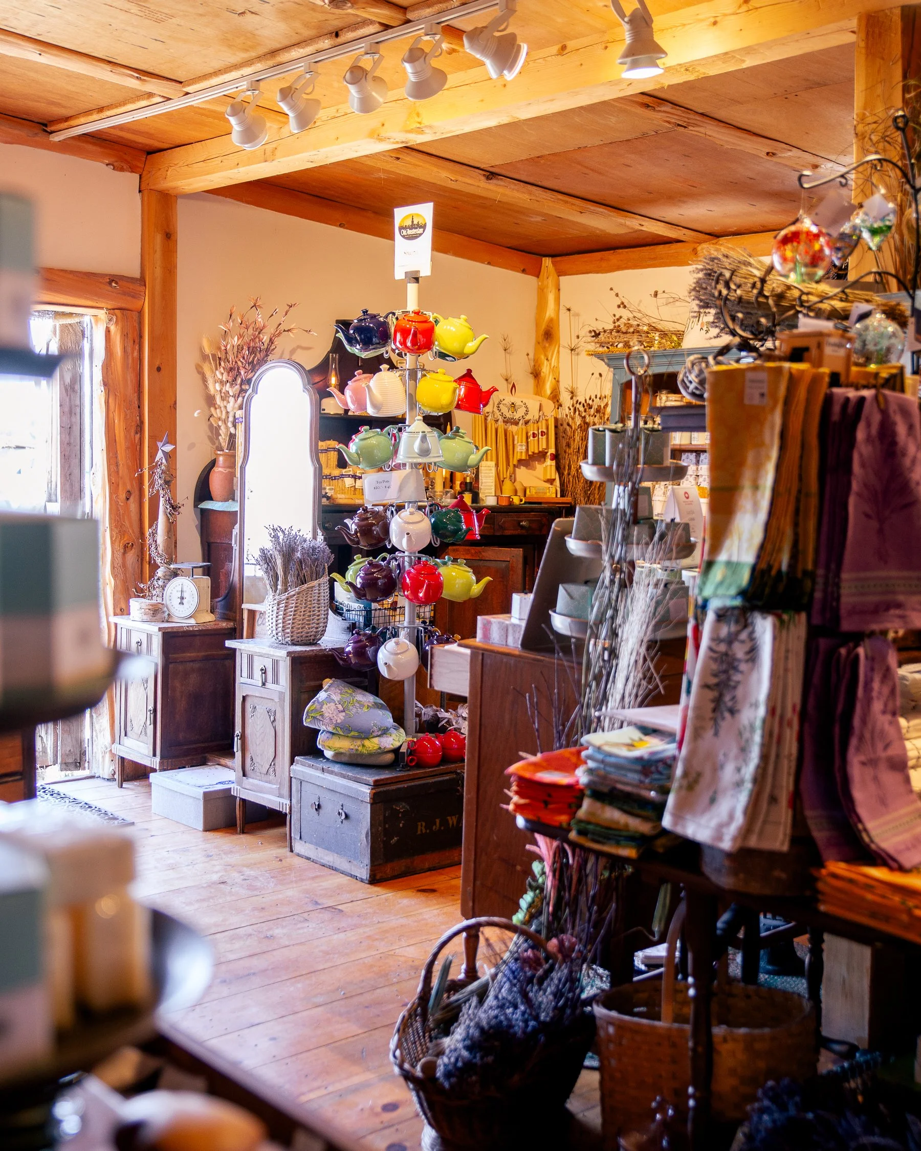 A cozy interior of a gift or home decor shop with wooden furniture, shelves, and a display of colorful teapots in the center. Many items like soaps, lotions, tea towels, and lavender bundles surround the shop