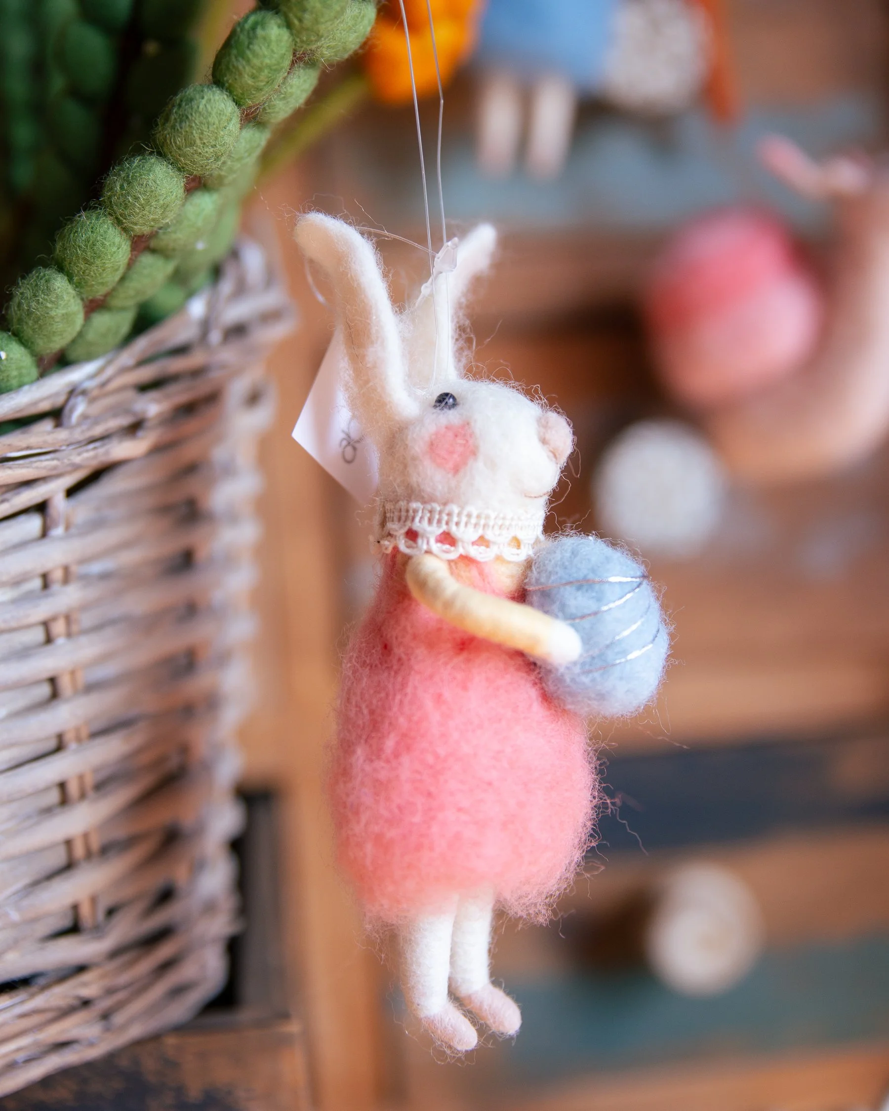 A hanging wool felt mouse ornament wearing a pink dress, holding a blue ball of yarn or egg, with a lace collar and pink cheeks, near a wicker basket with green plants. More felt animals blurred in the background