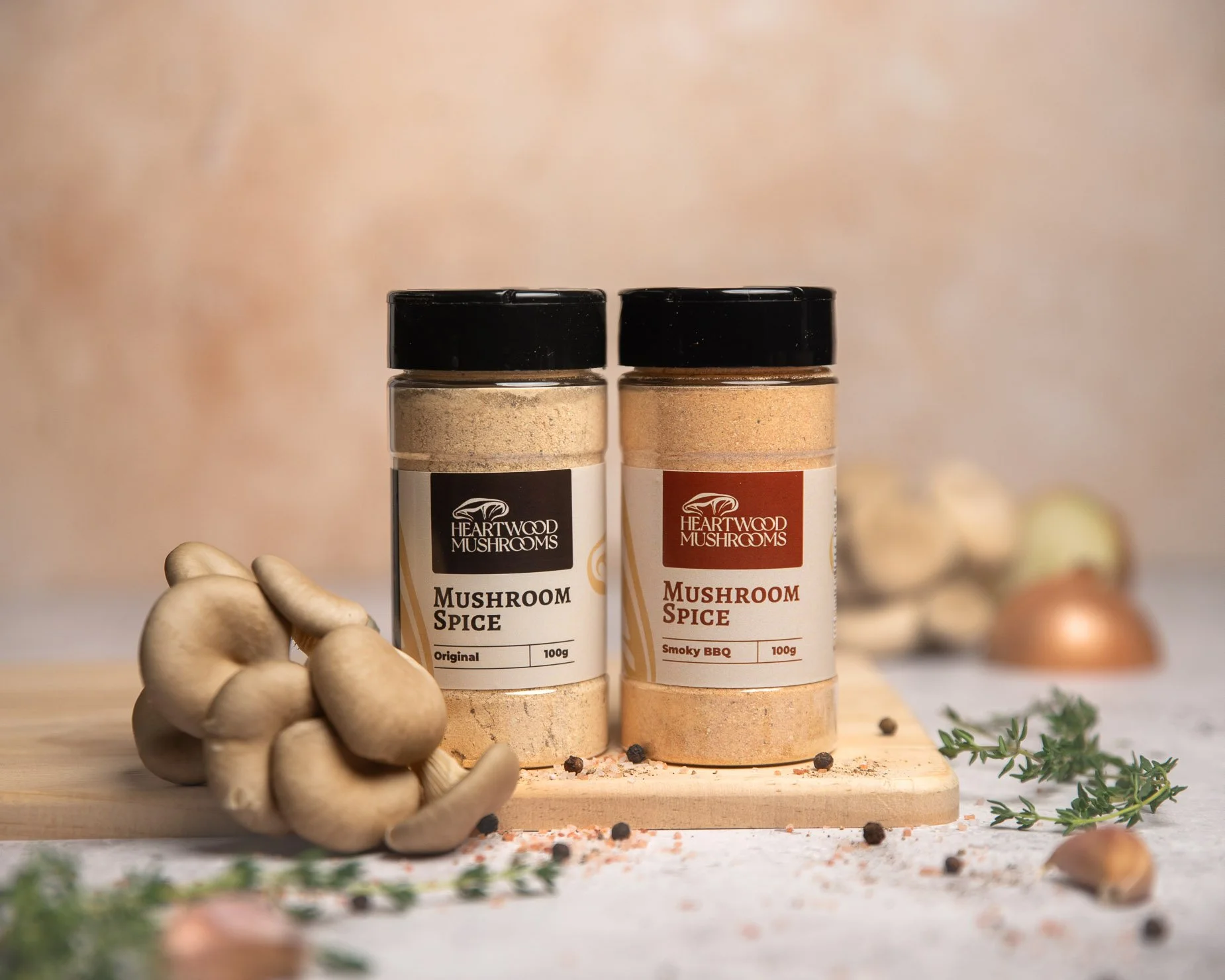 Two jars of mushroom spice from Heartwood Mushrooms, one labeled Original and the other Smoky BBQ, placed on a wooden surface surrounded by fresh mushrooms, herbs, peppercorns, and garlic.