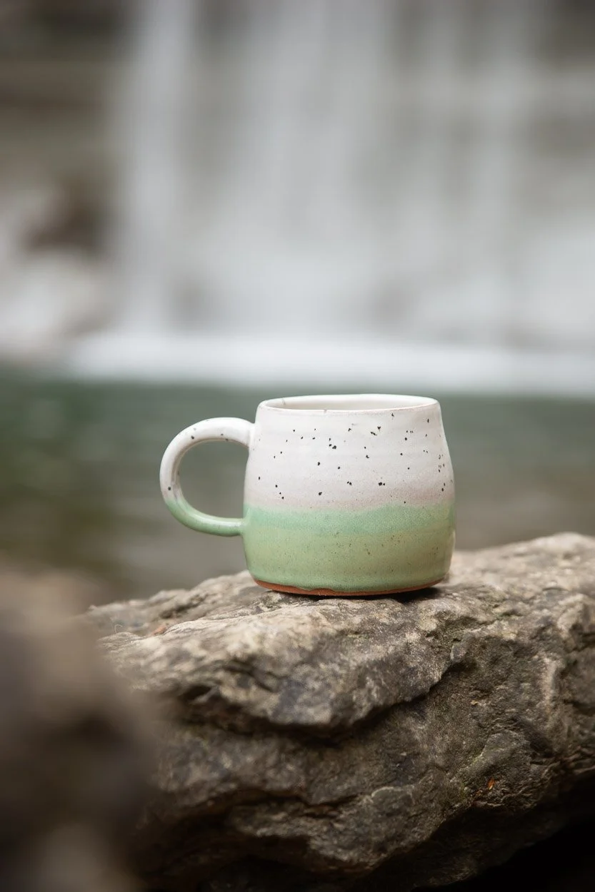 A ceramic mug with a speckled white and green glaze rests on a large rock near a waterfall with a rock in the foreground