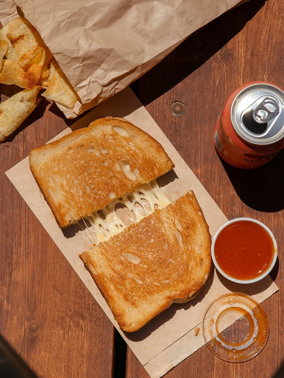 Grilled cheese sandwich with melted cheese, potato chips in a paper bag, a can of soda, and a small dipping cup on homemade tomato ketchup on a wooden table.