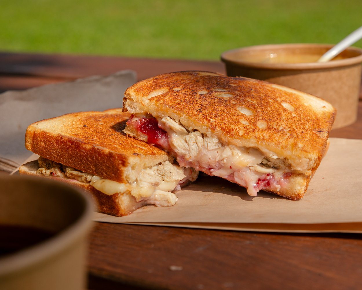 A toasted grilled cheese sandwich with turkey and cranberry sauce inside, cut in half, on a piece of parchment paper. In the background, there's a bowl of soup and a small cup of dipping sauce, on a wooden table outdoors.