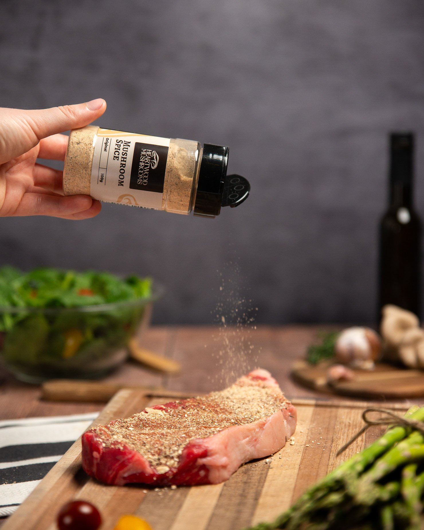 Hand sprinkling Heartwood Mushrooms' mushroom spice over a raw steak on a wooden cutting board with vegetables and cooking ingredients in the background, a striped tea towel under the cutting board, and asparagus in the foreground