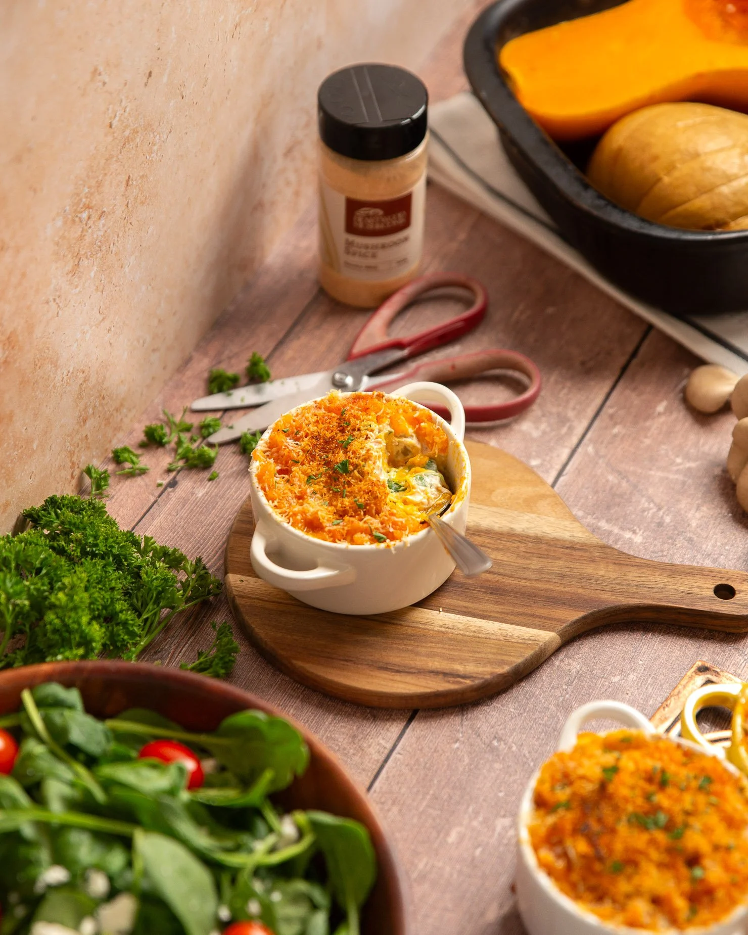 Baked casserole in a white ramekin on a wooden serving board, garnished with herbs, on a rustic wooden table with fresh vegetables and kitchen tools nearby. A jar of 'smoky BBQ' mushroom spice from Heartwood Mushrooms is blurred in the background