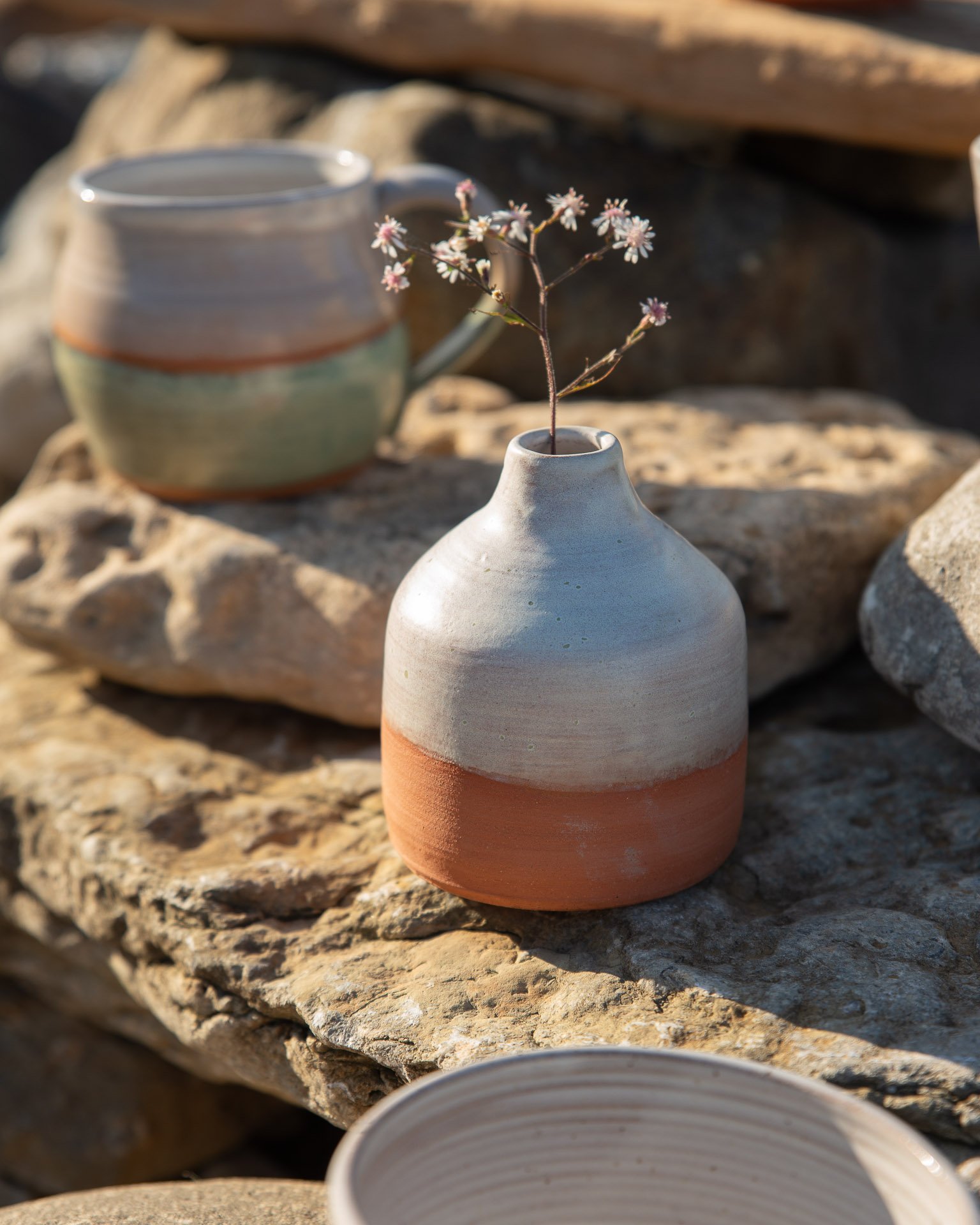 A small ceramic vase with a two-tone glaze, containing a delicate sprig of small pink flowers, placed on a rocky surface outdoors with more ceramic items in the foreground and background