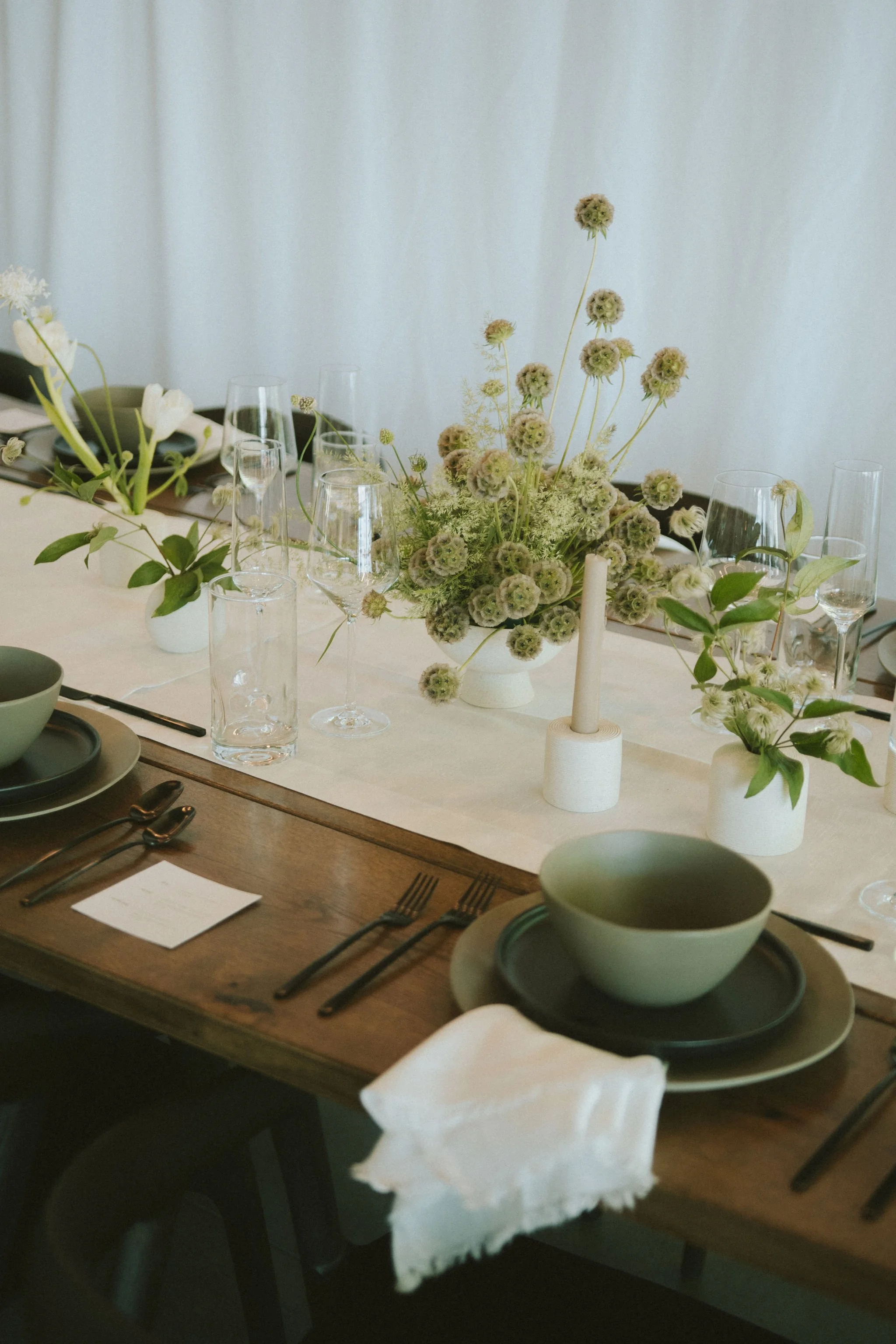 A dining table set with green bowls and plates, black utensils, clear glasses, and small white vases with green and white flowers and greenery, with a white candle in a white holder and a beige table runner.