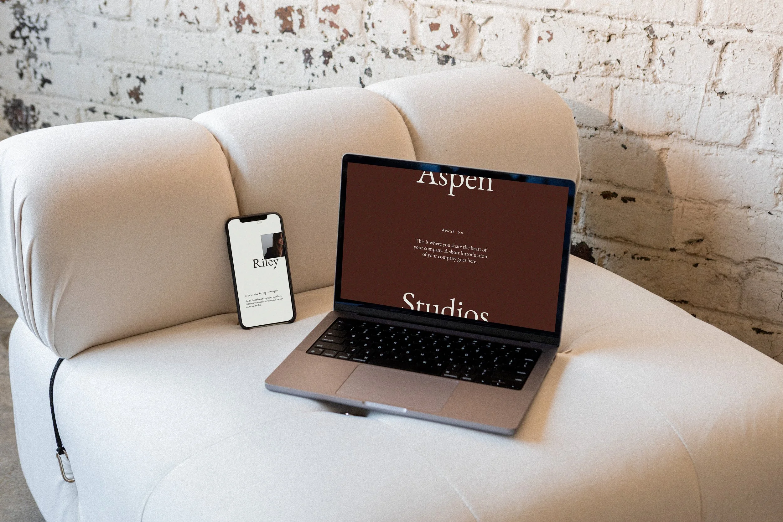 A white upholstered sofa with a laptop and a smartphone resting on it, against a white brick wall. The laptop screen displays 'Aspen Studios'. The smartphone screen shows a person named Riley with some text below.