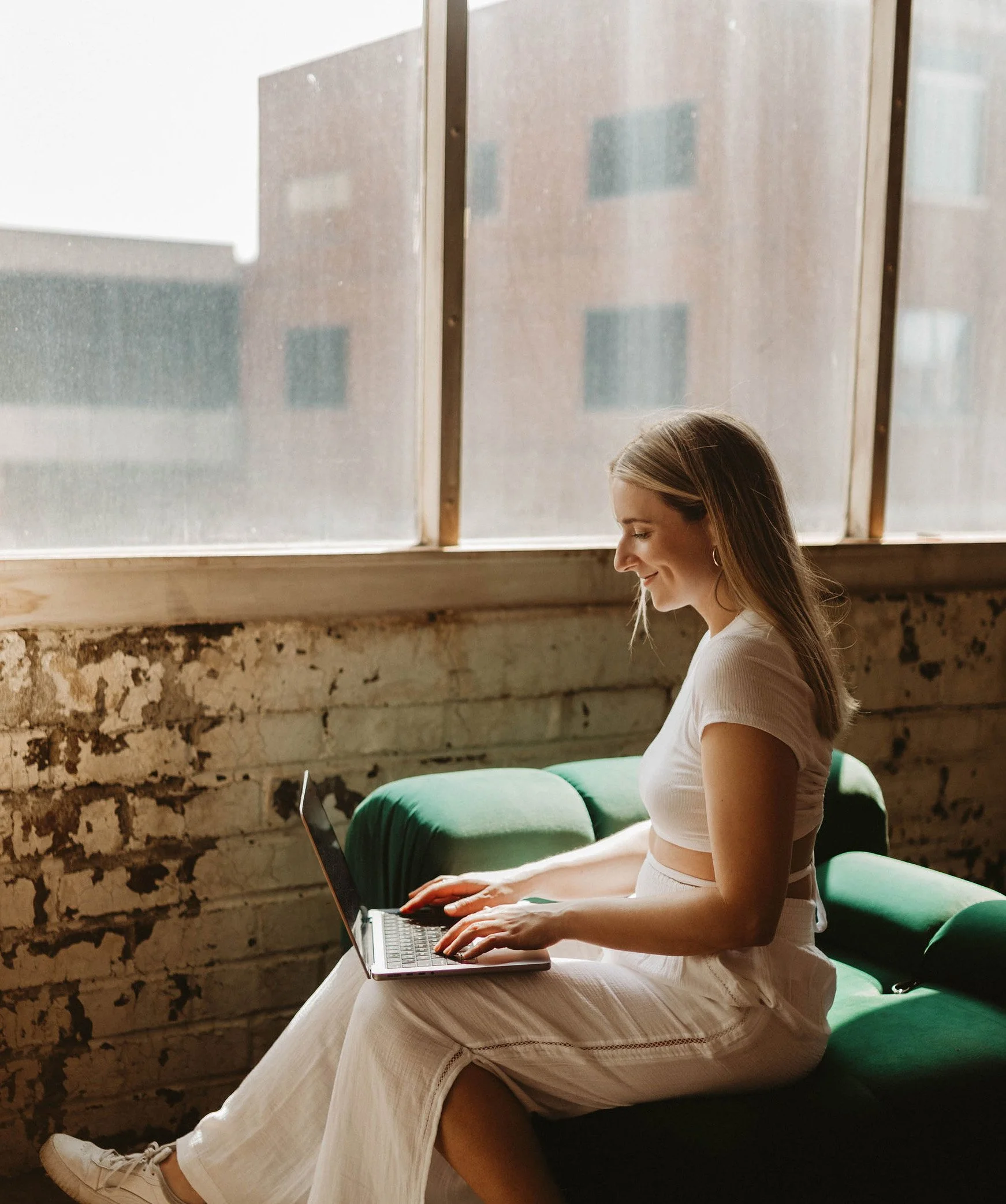 A young woman sitting on a green couch working on a laptop, near a large window with a cityscape outside, in a cozy, rustic room.