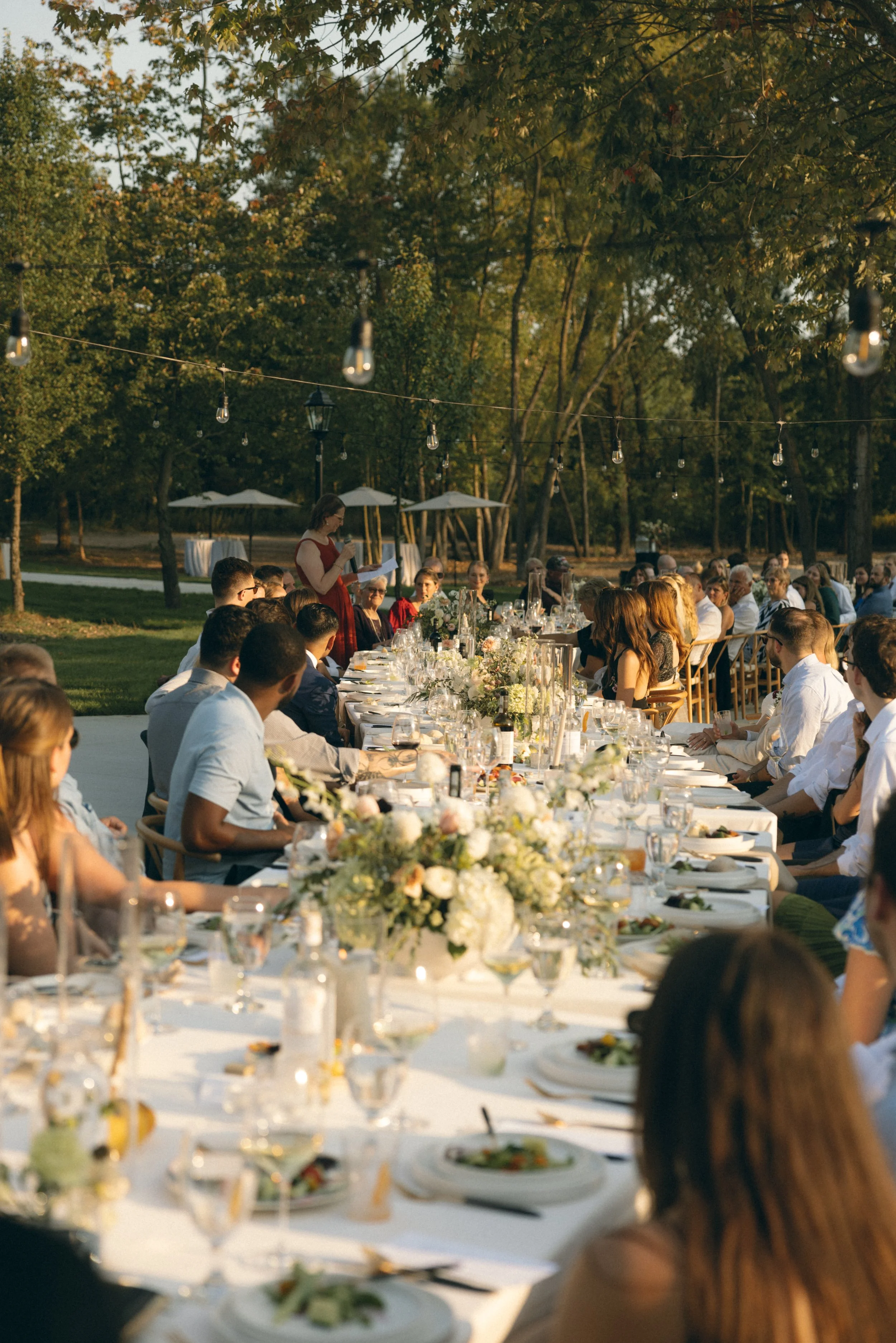 People seated at arranged outdoor wedding reception dining table decorated with flowers and elegant tableware during sunset.