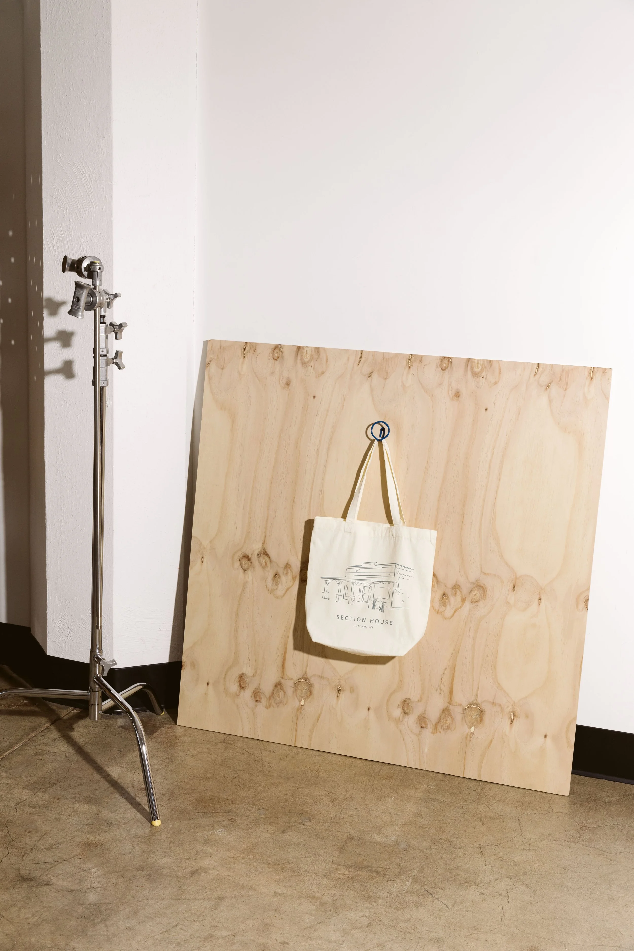 A tote bag with a line drawing of a building hangs on a hook on a plywood board leaning against a white wall. To the left, there is a tall studio light stand with adjustable knobs.