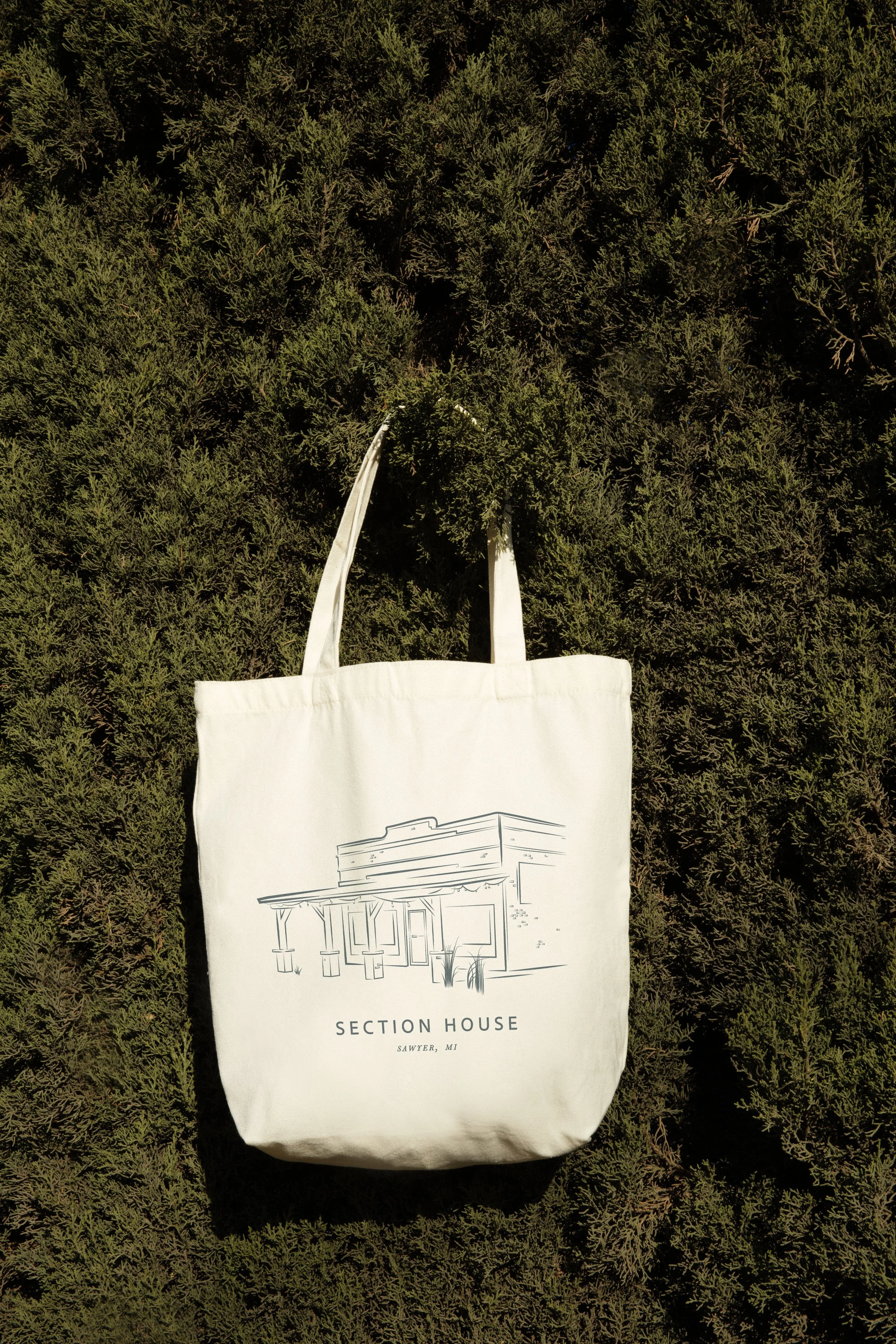 A white tote bag with a line drawing of a building and the words 'Section House, Sawyer, MI' printed on it, laying on a bed of green shrubbery.