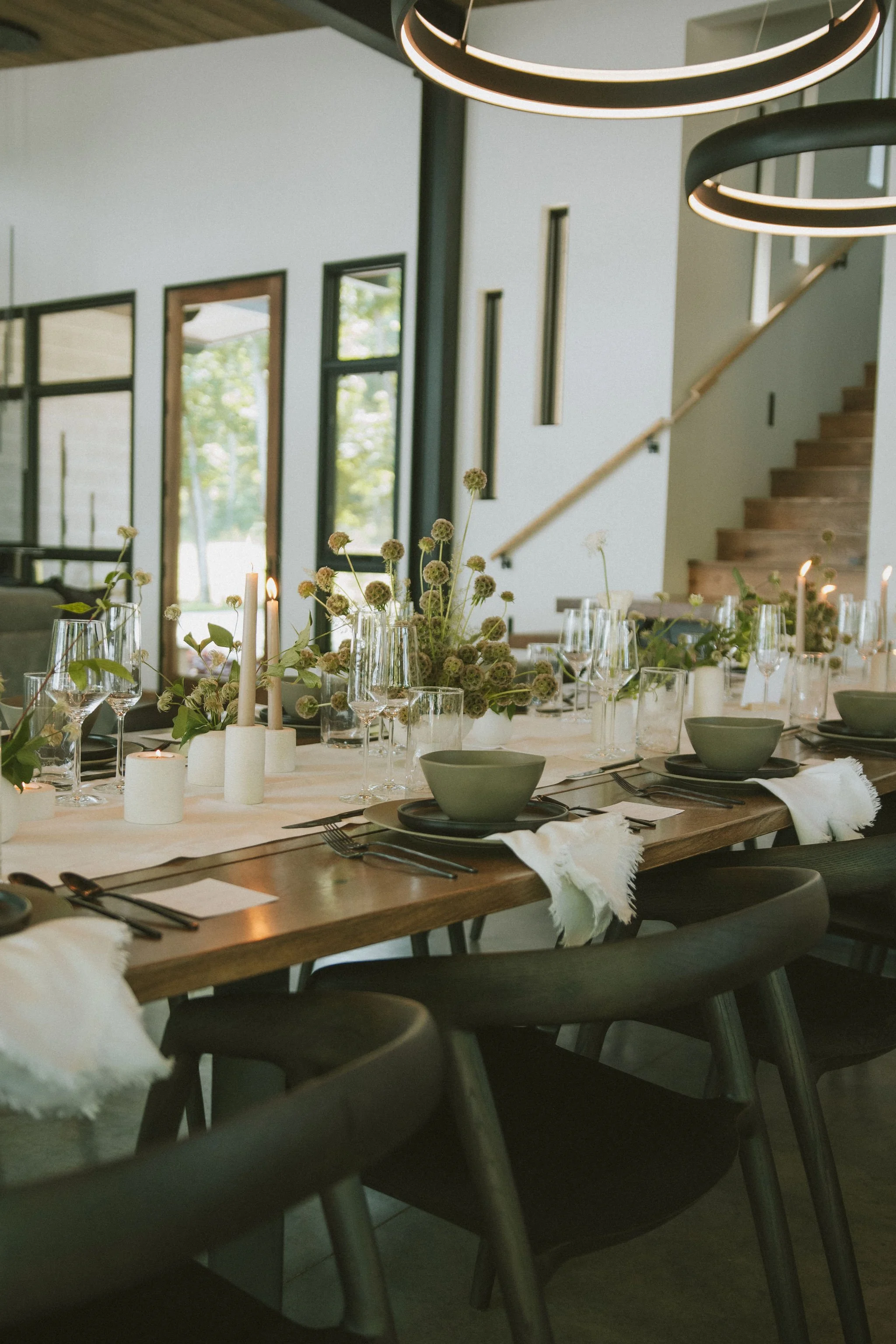 A lively dining room set for a formal dinner with a long wooden table, surrounded by black chairs, decorated with candles, greenery, and elegant tableware, in a modern house with large windows and a staircase.