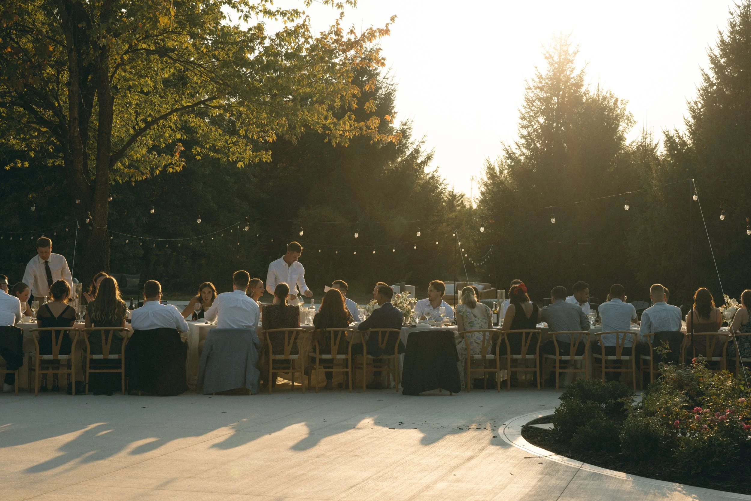 People sitting at a long outdoor banquet table during sunset in a lush garden, with string lights hanging overhead.