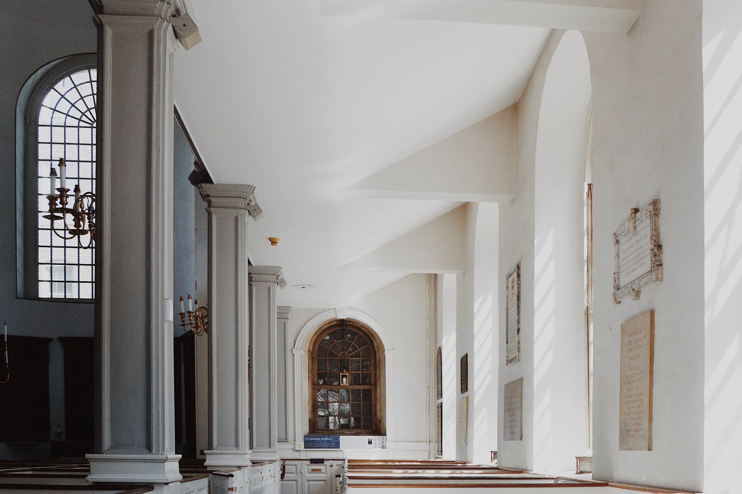 Interior of a church with tall white columns, arched windows, and sunlight streaming through narrow windows on the right side, creating shadows on the wall.