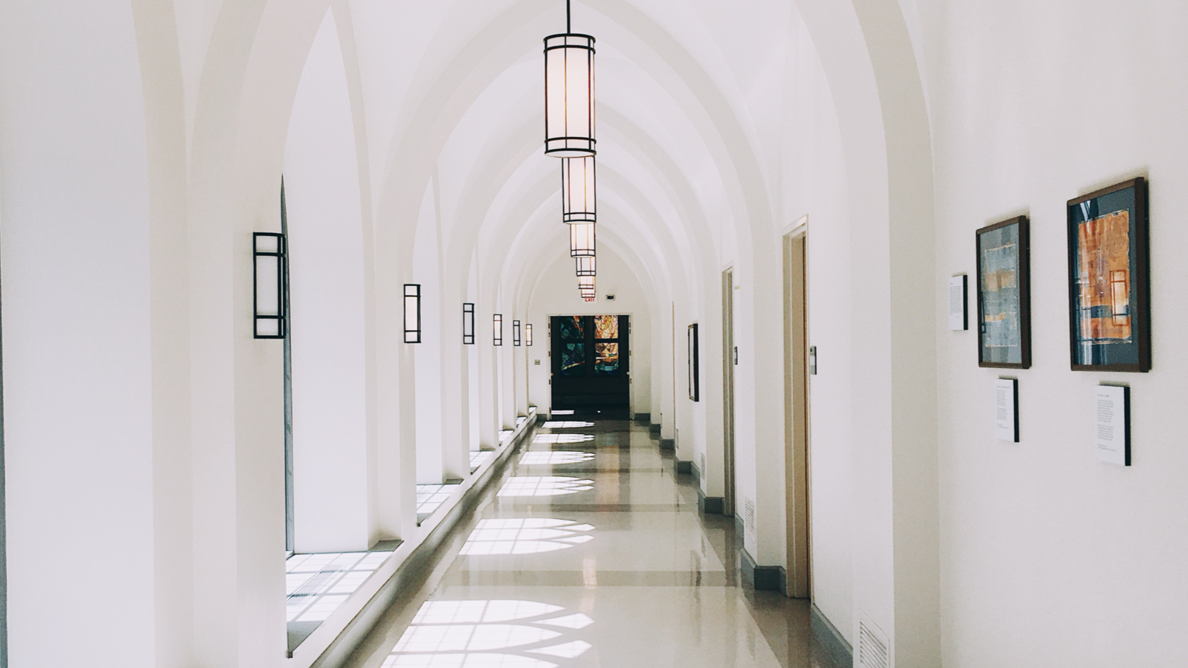 A bright, arched hallway with hanging light fixtures and framed artwork on the wall, sunlight casting shadows on the floor.
