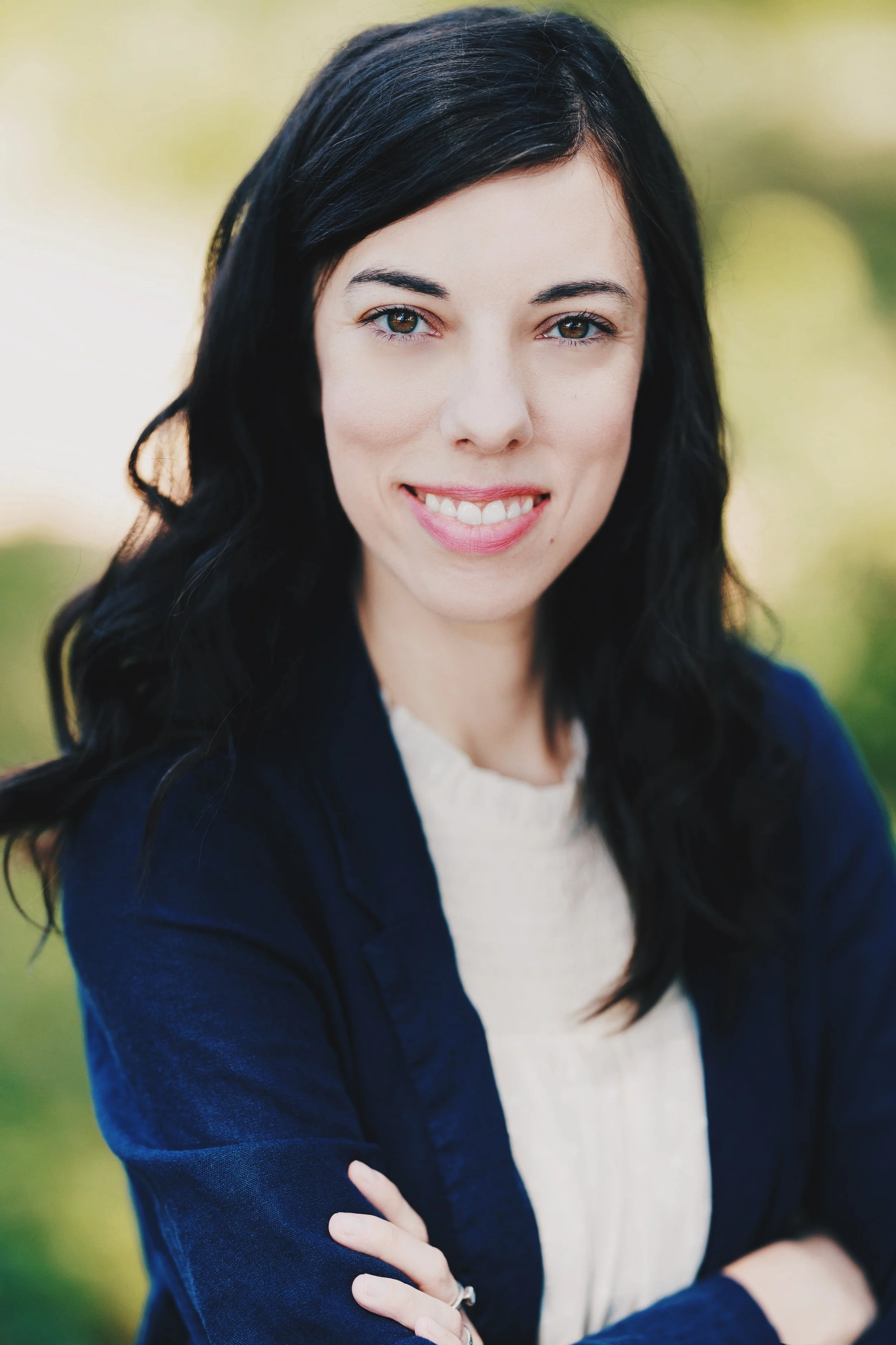 A portrait of a woman with long black hair, smiling, wearing a navy blue blazer and a light-colored top, outside with blurred greenery in the background.