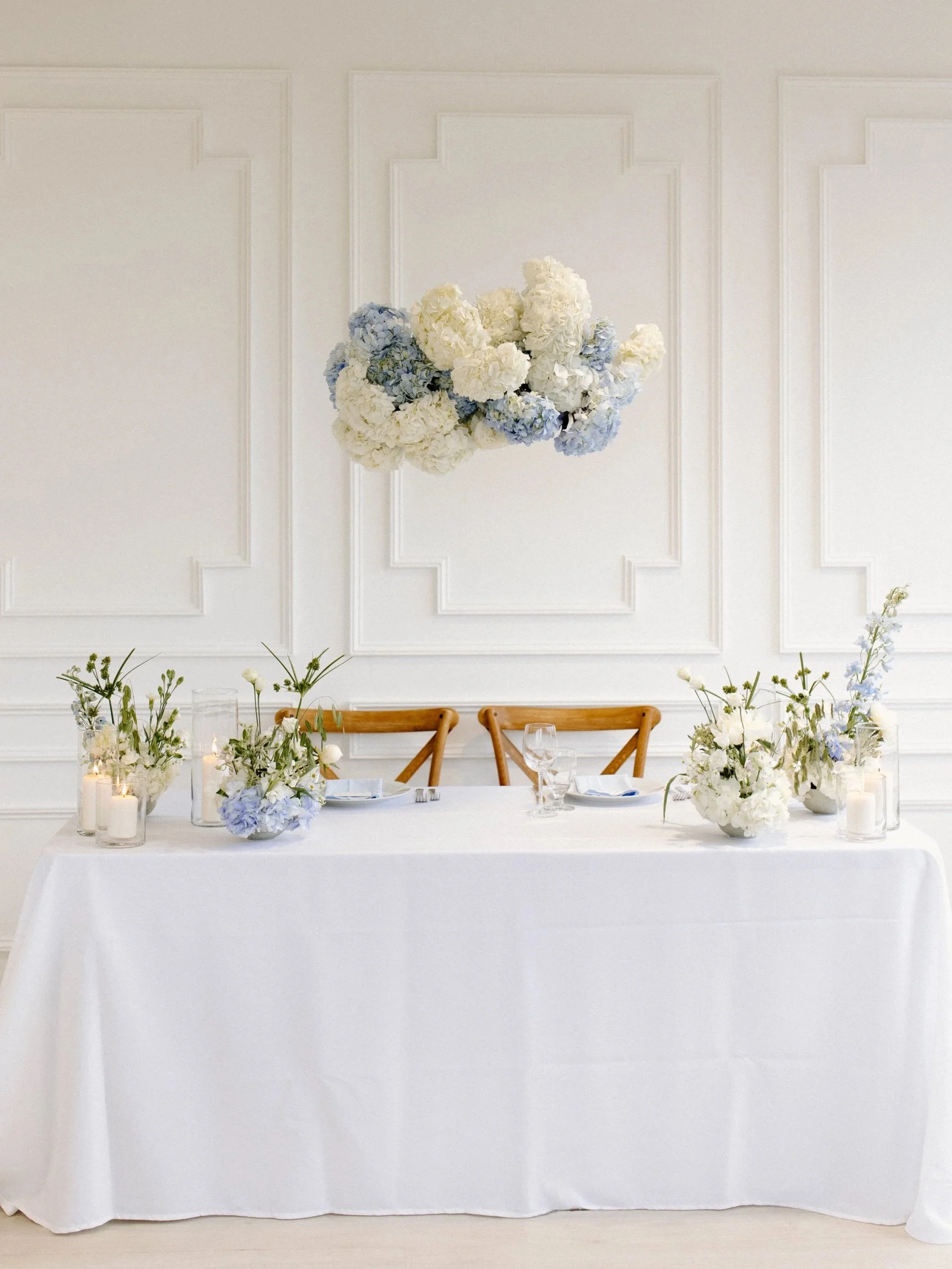 Elegant table setup with white tablecloth, floral arrangements with white and light blue flowers, candles, and place settings, against a white paneled wall with a large hanging floral arrangement of white and light blue hydrangeas.