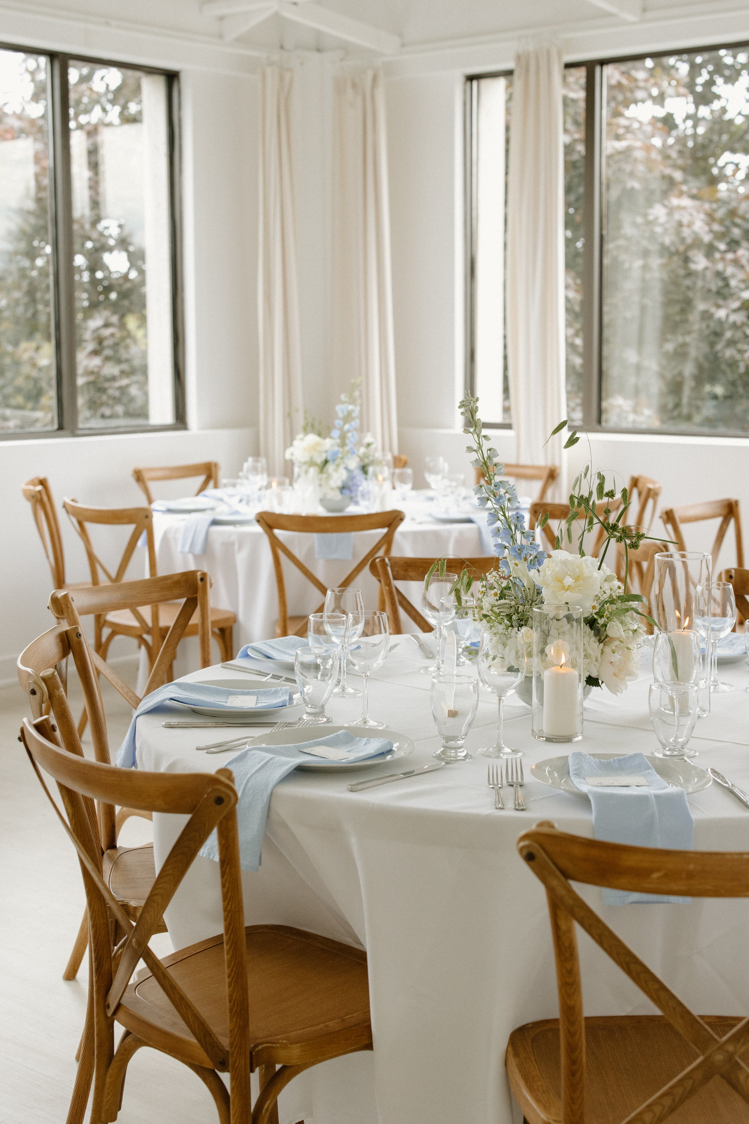 A decorated round banquet table set for a formal event with white tablecloths, blue napkins, glassware, silverware, and a floral centerpiece with white and blue flowers, candles, and greenery, in a naturally lit room with large windows and white curtains.