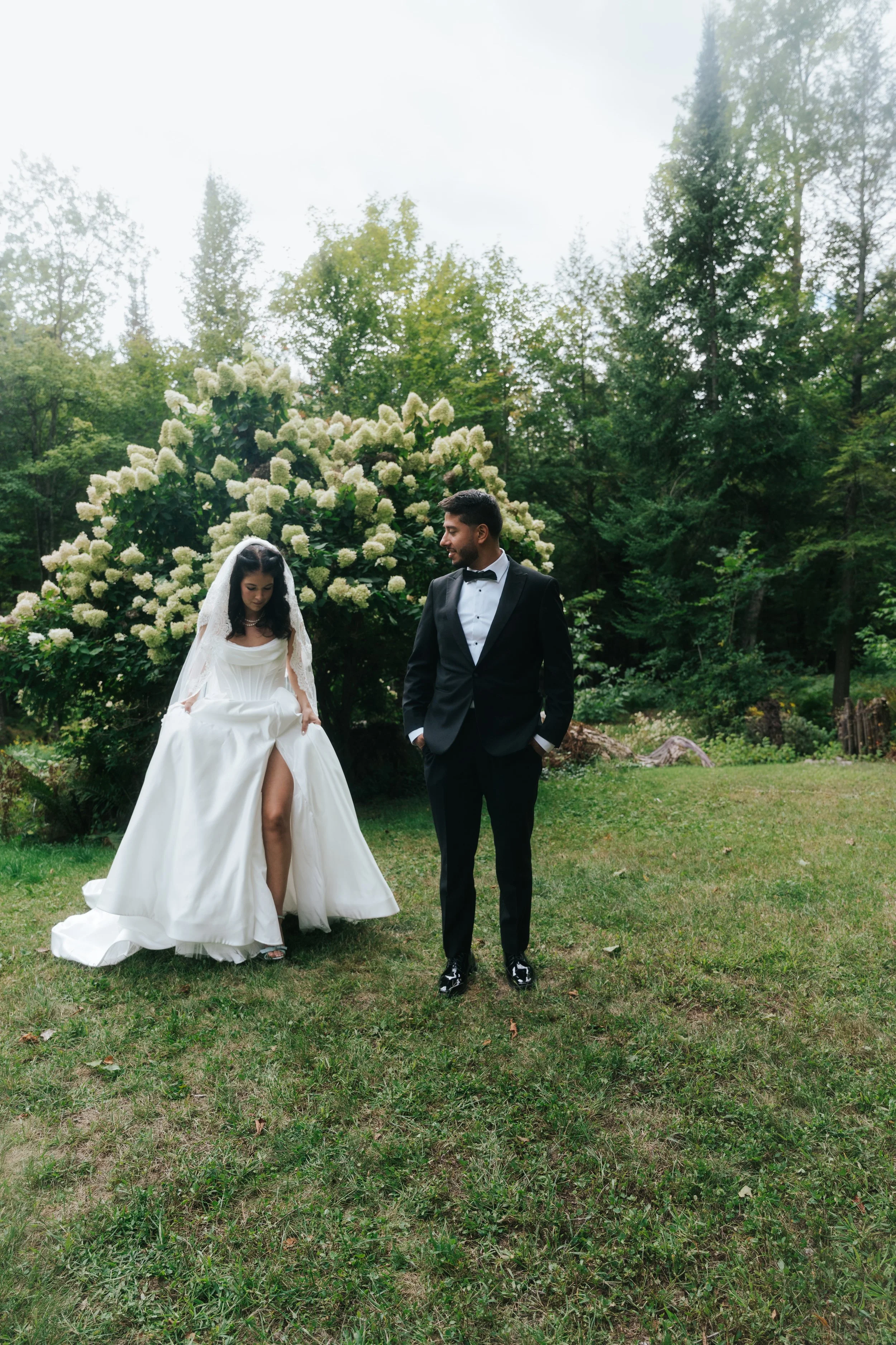 A bride in a white wedding dress lifting her skirt to reveal her legs, standing next to a groom in a black tuxedo with a bow tie, outdoors on a grassy area with green trees and a large flowering bush in the background.
