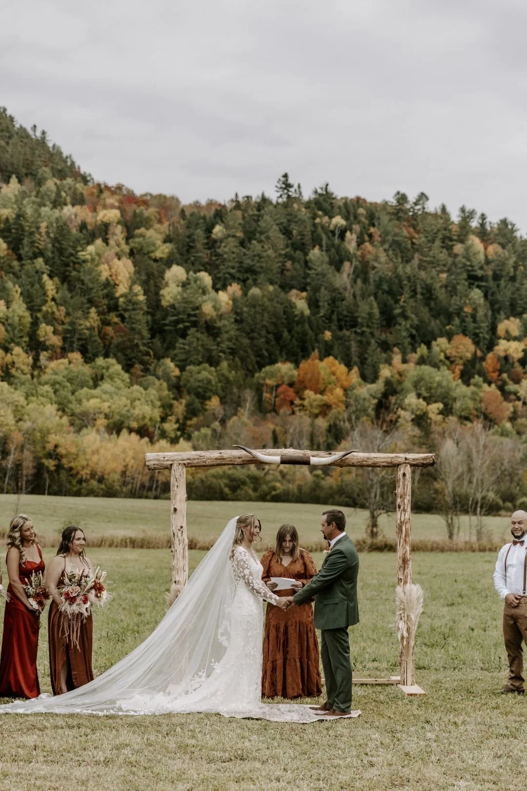 A wedding ceremony outdoors in a grassy field with a mountain backdrop. The bride in a white lace dress and long veil and the groom in a green suit hold hands under a rustic wooden arch. Bridesmaids and groomsmen, dressed in rust-colored and white outfits, stand nearby.