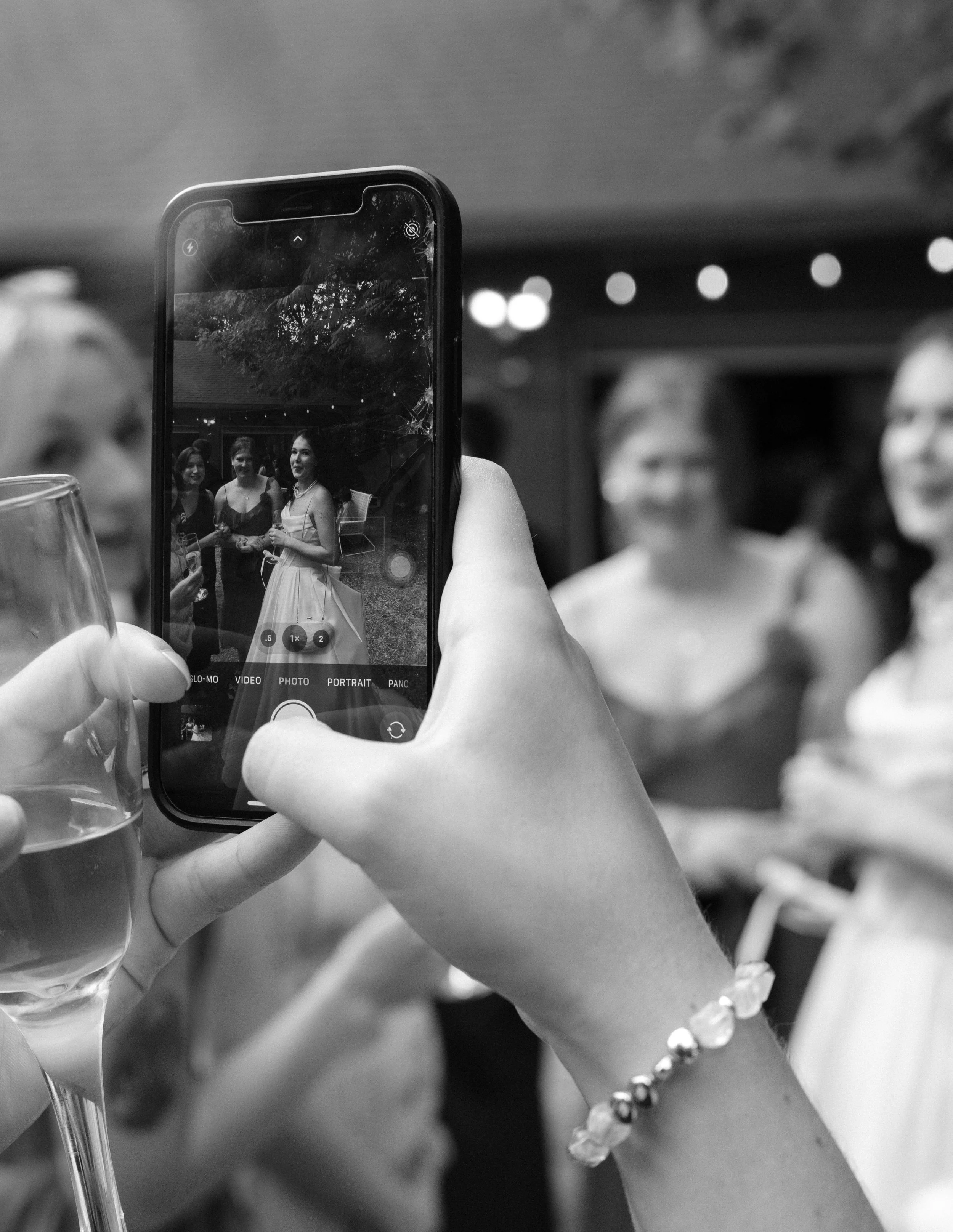 A person taking a photo on their phone of a group of women at a social gathering. The women are smiling and holding drinks, with blurred background lights.