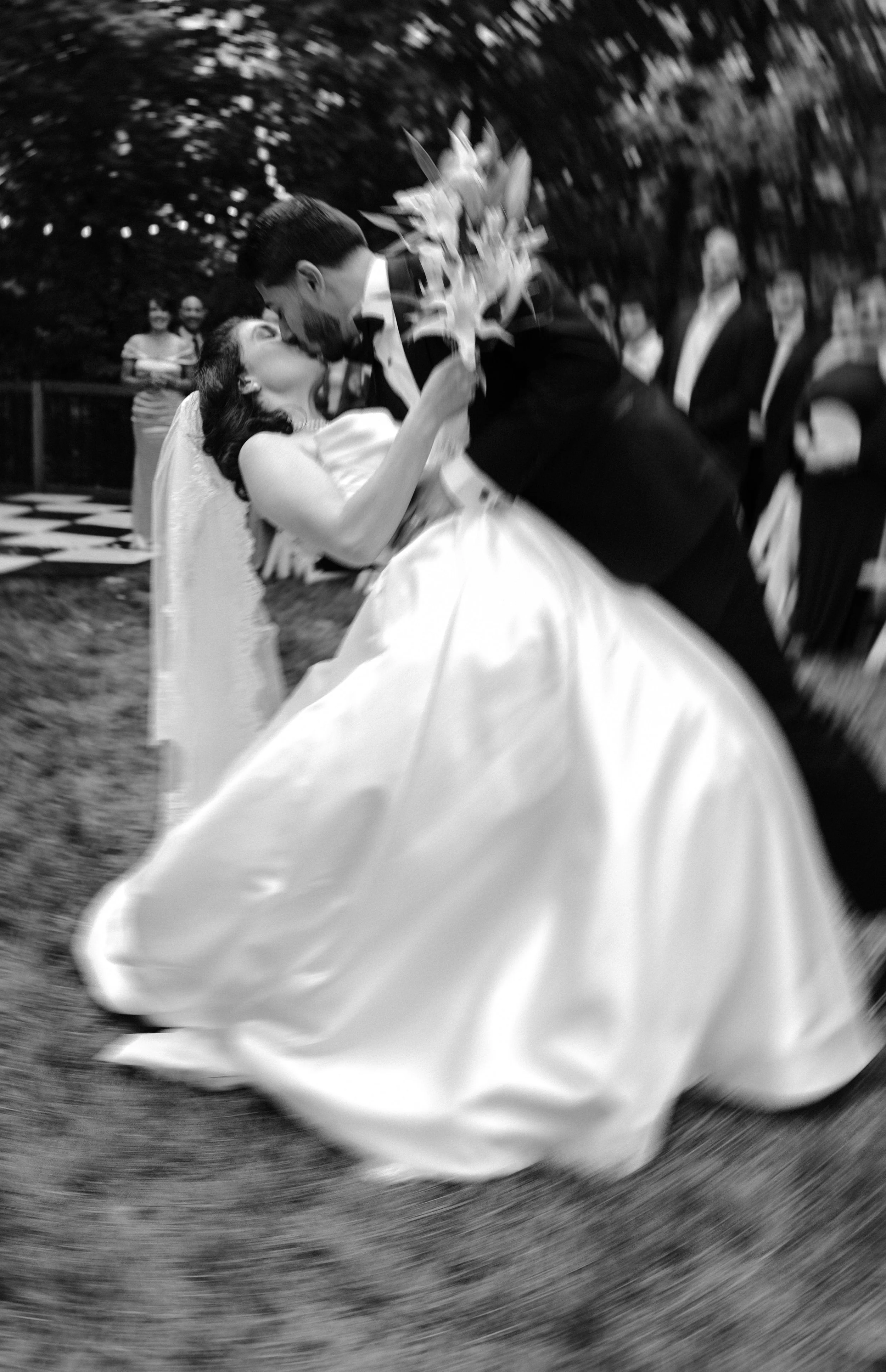 A bride and groom sharing a kiss during their wedding ceremony outdoors, with friends and family watching in the background.