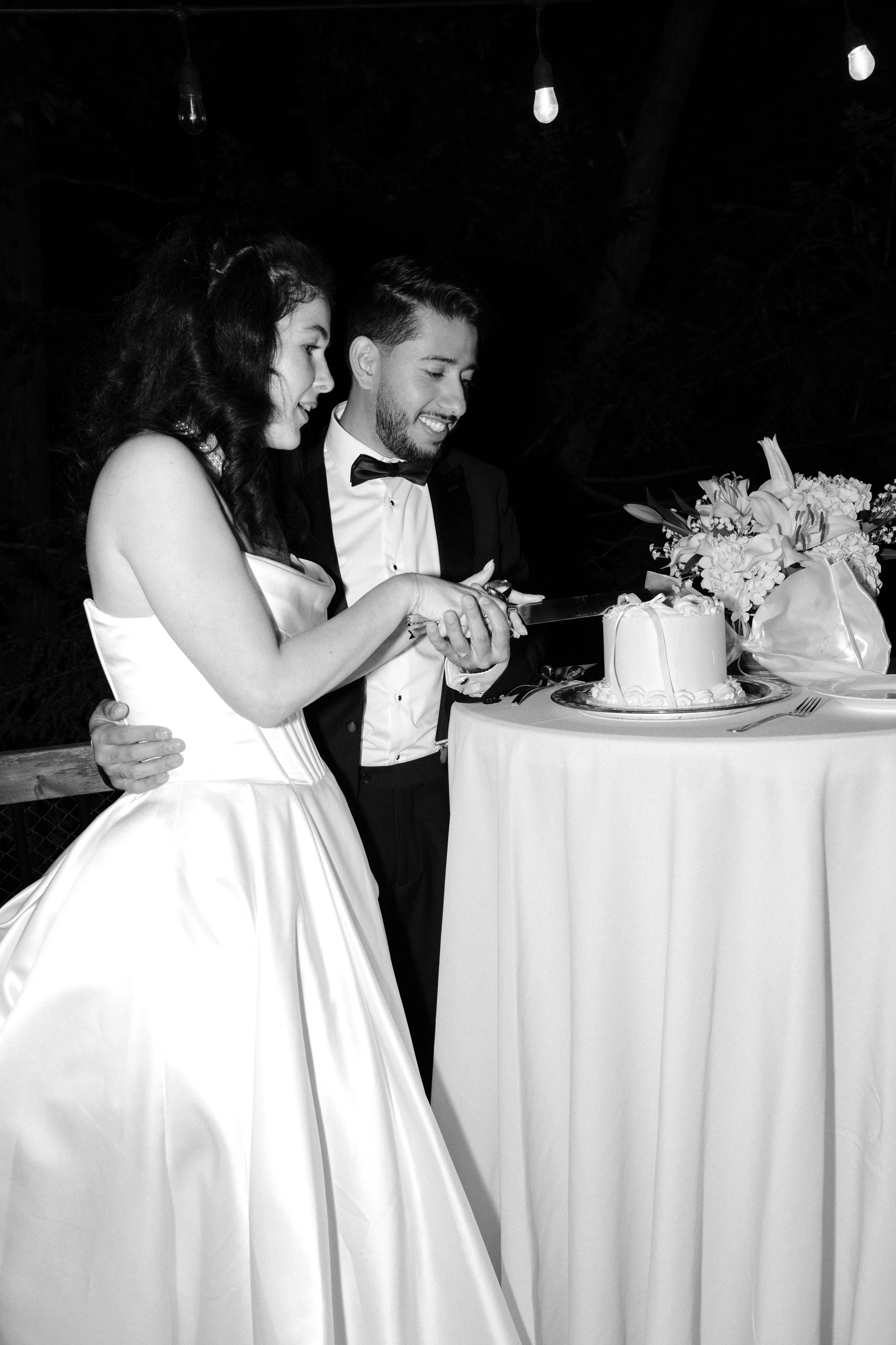 A bride and groom cutting a wedding cake outdoors at night, with string lights overhead.
