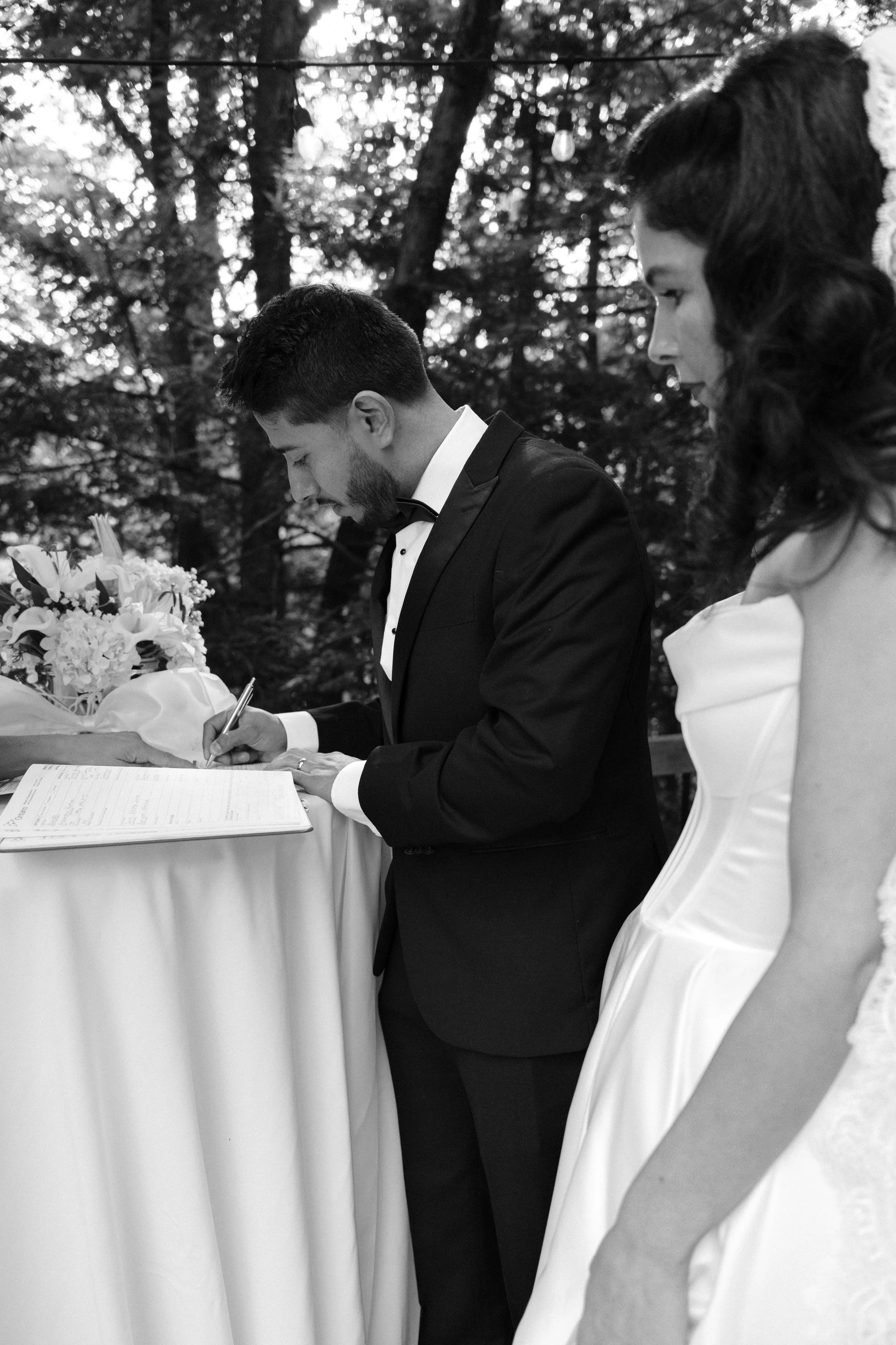 A black-and-white photo of a groom signing a marriage certificate outdoors with trees in the background. The bride is partially visible standing beside him.