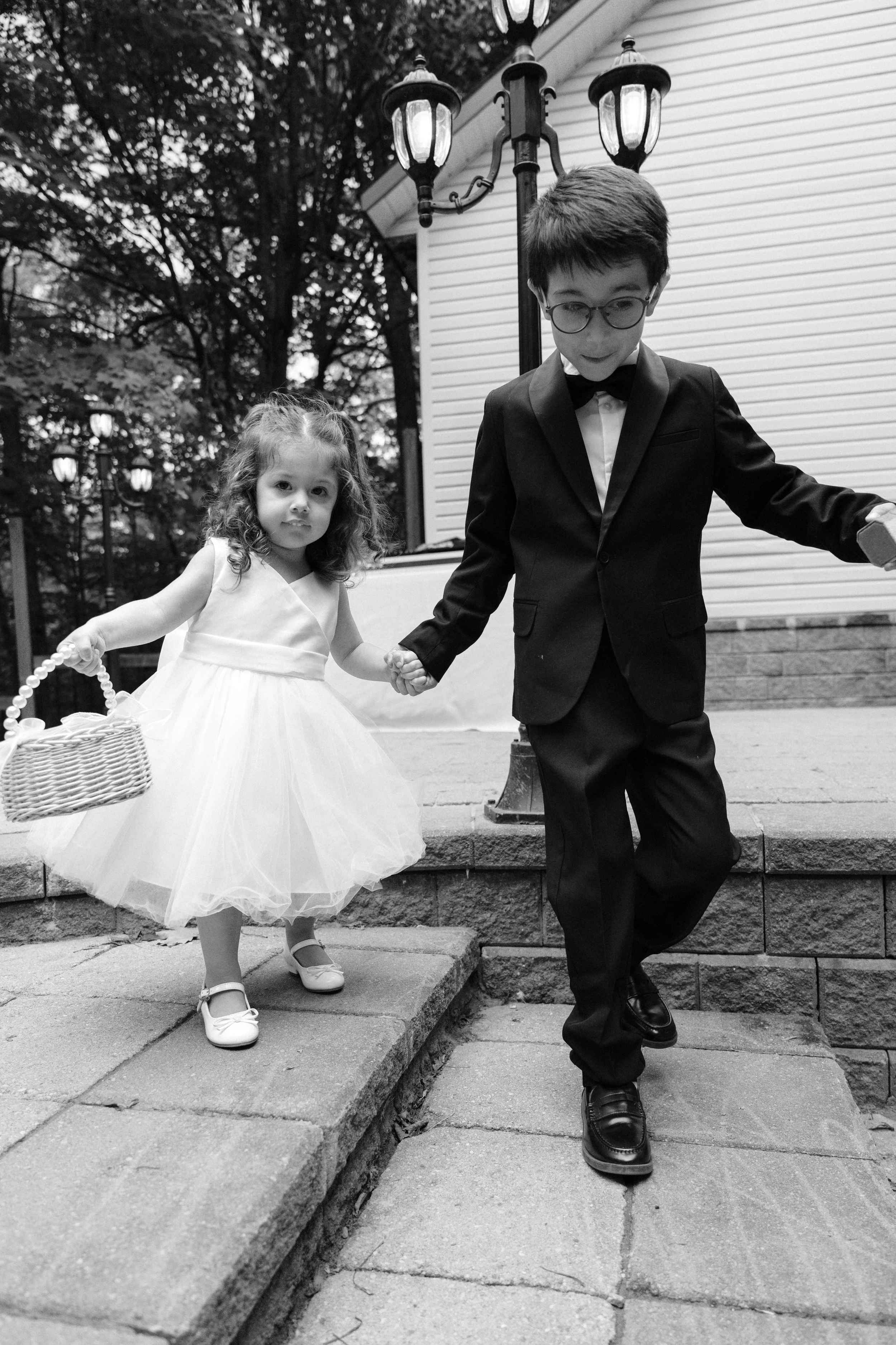A young boy and girl, dressed in formal attire, walking down outdoor stone steps holding hands. The girl is wearing a white dress and holding a basket, while the boy is wearing a suit with a bow tie. There are trees and street lamps in the background