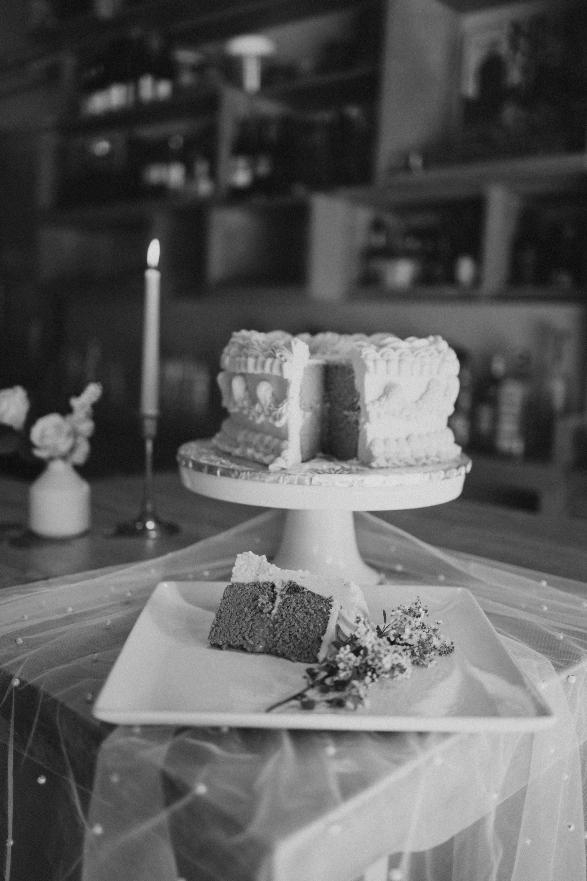 A slice of cake on a white plate with flowers, placed on a table covered with a sheer cloth. Behind it is a larger cake on a cake stand. In the background, there is a candle and a small vase with flowers.