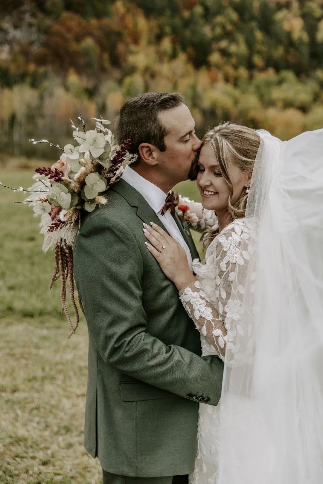 A newlywed couple embracing outdoors during autumn. The groom, in a green suit, kisses the bride, who is wearing a lace wedding dress. She is smiling, and he has a bouquet with white and dried flowers on his back.