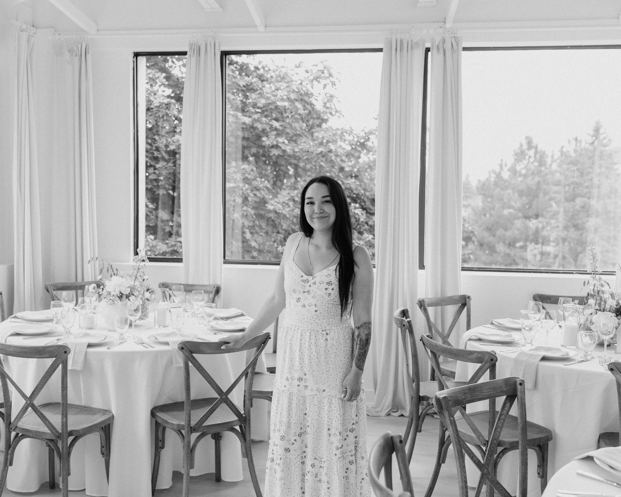 A woman with long dark hair wearing a white dress stands in a dining room with round tables set for a meal, decorated with flowers, plates, glasses, and silverware, near large windows with curtains, during daytime.