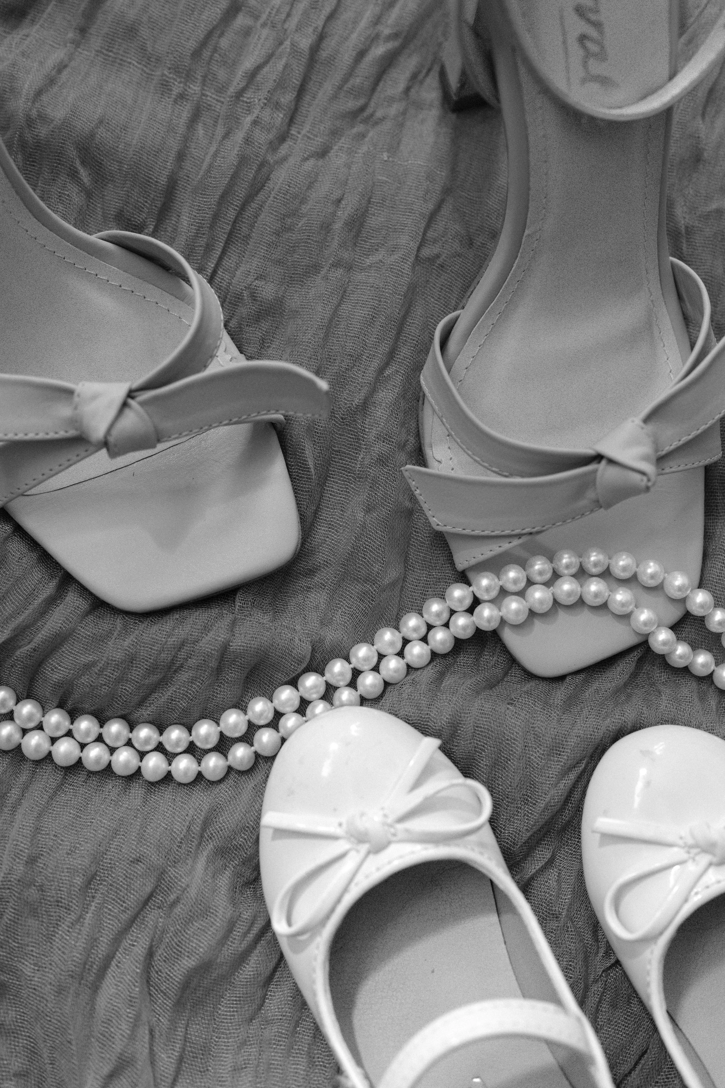 Pair of white high-heeled shoes with bows, pearl necklace, and a milk jug on a textured fabric surface.