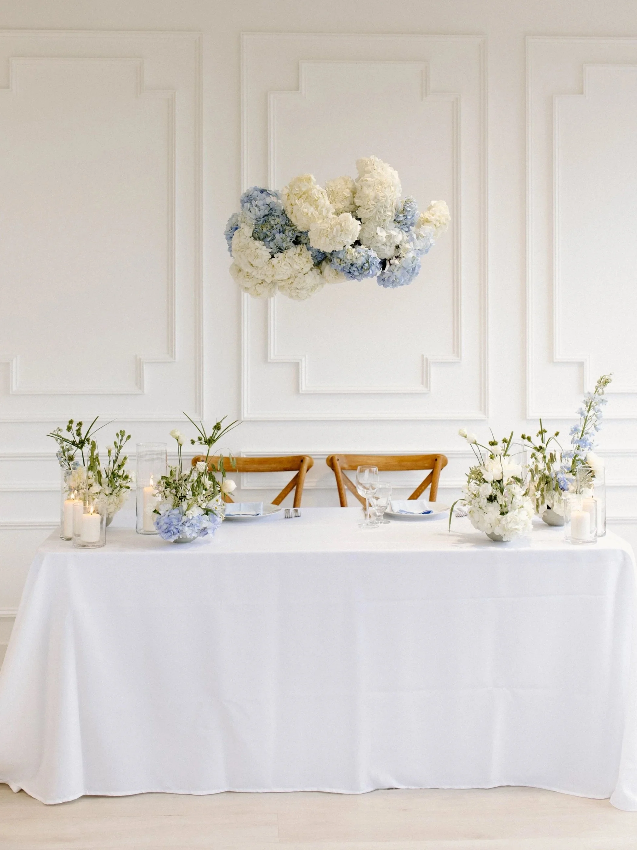 Elegant table setup with white tablecloth, floral centerpieces, and candles, featuring a lush flower arrangement of white and blue hydrangeas hanging on the white paneled wall behind.
