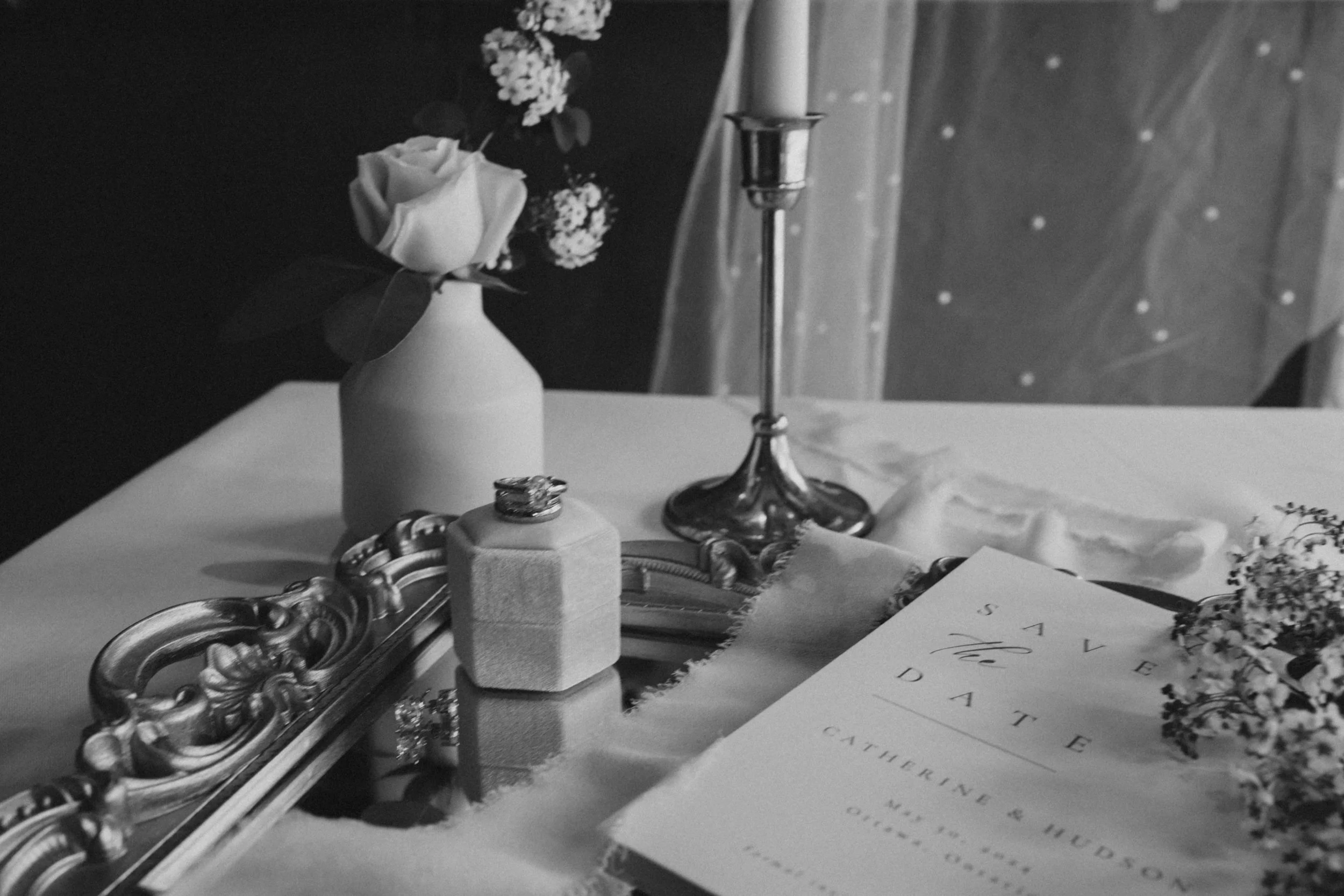 A black and white photograph of a wedding table setting, including a small bouquet of roses and flowers in a vase, a candlestick, a mirror tray with jewelry, a ring, and a book titled 'Save the Date' for a wedding with details of the couple and wedding date.