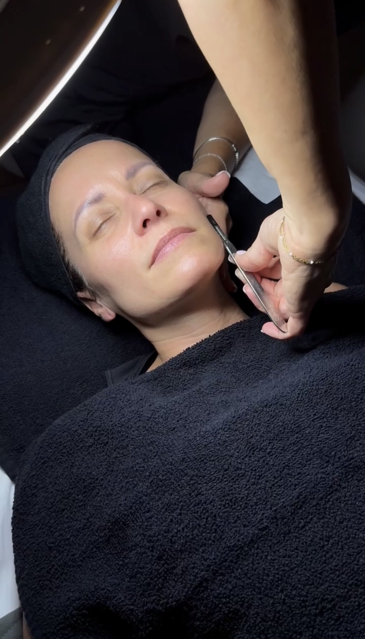 A woman receiving a facial treatment, lying down with her eyes closed, a towel wrapped around her head, while a professional uses a tool on her face.