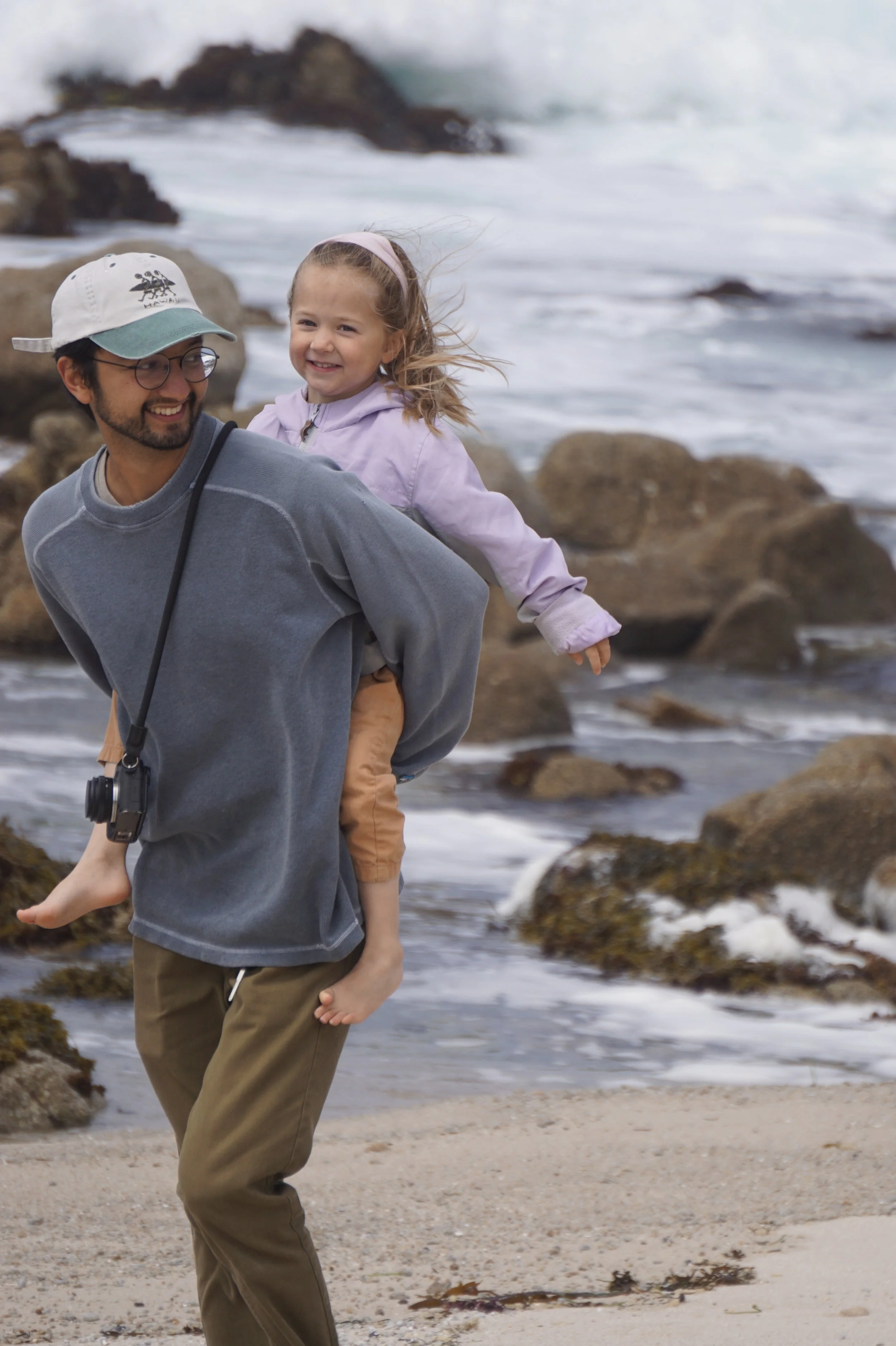 A man carrying a young girl on his back while walking on a beach with rocks and waves in the background.