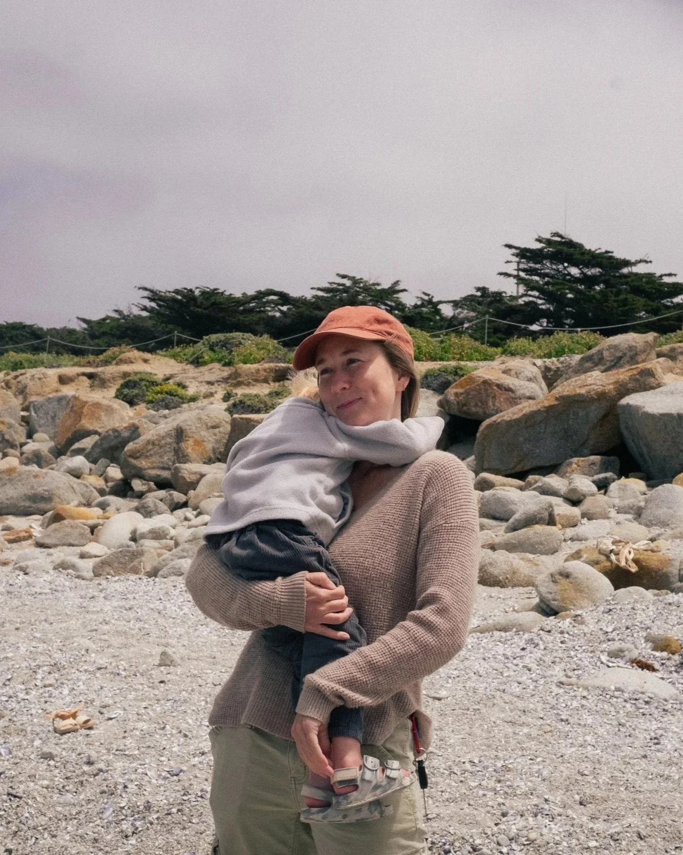 A woman with a brown cap and beige sweater holding a young boy in gray hoodie and dark pants on a rocky beach with large rocks and trees in the background.
