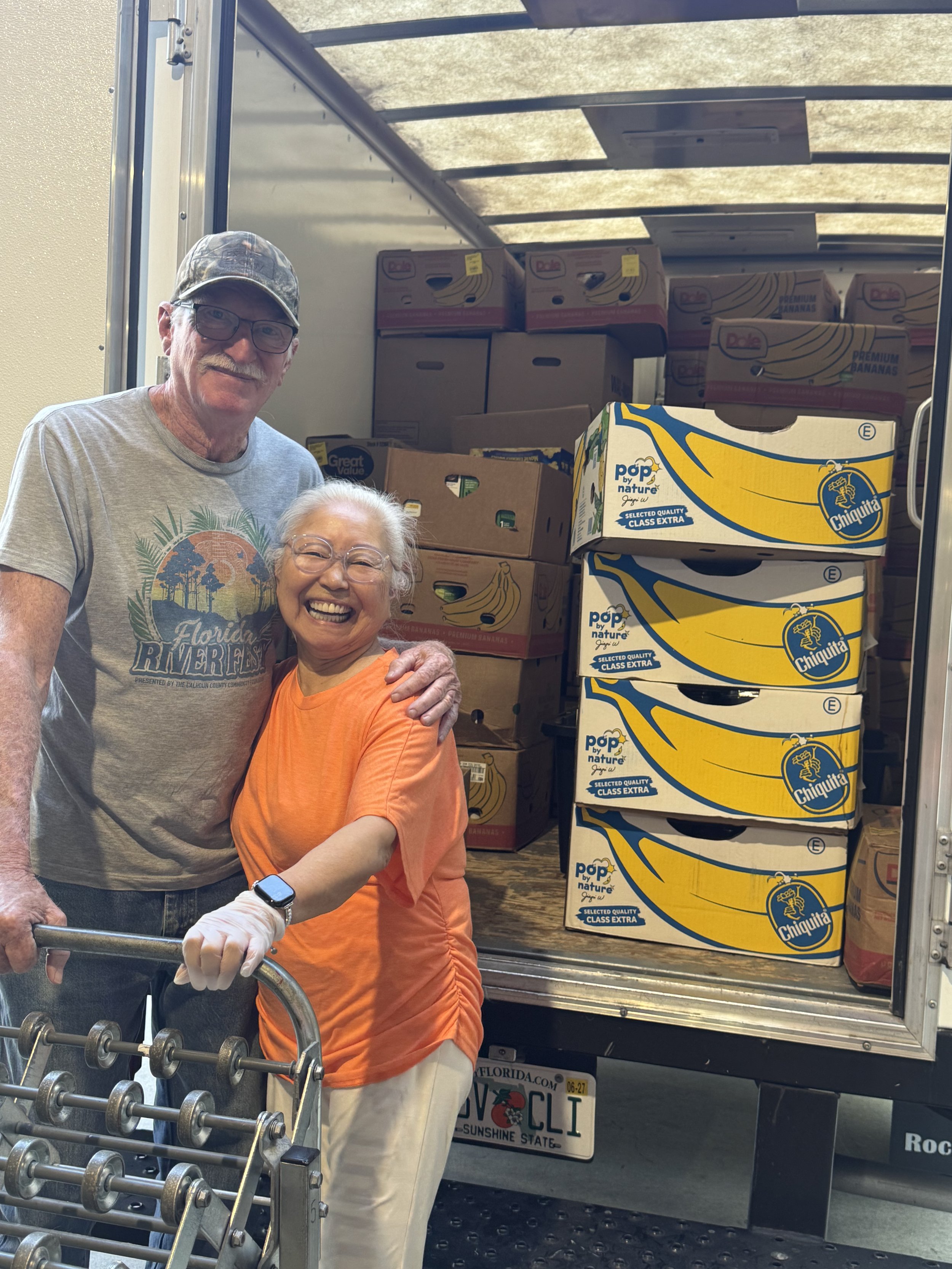 An elderly woman smiling and holding onto a shopping cart, in front of a truck loaded with boxes of bananas and other groceries. A man stands beside her, wearing a camo cap, glasses, and a grey t-shirt. The truck has a Florida license plate.